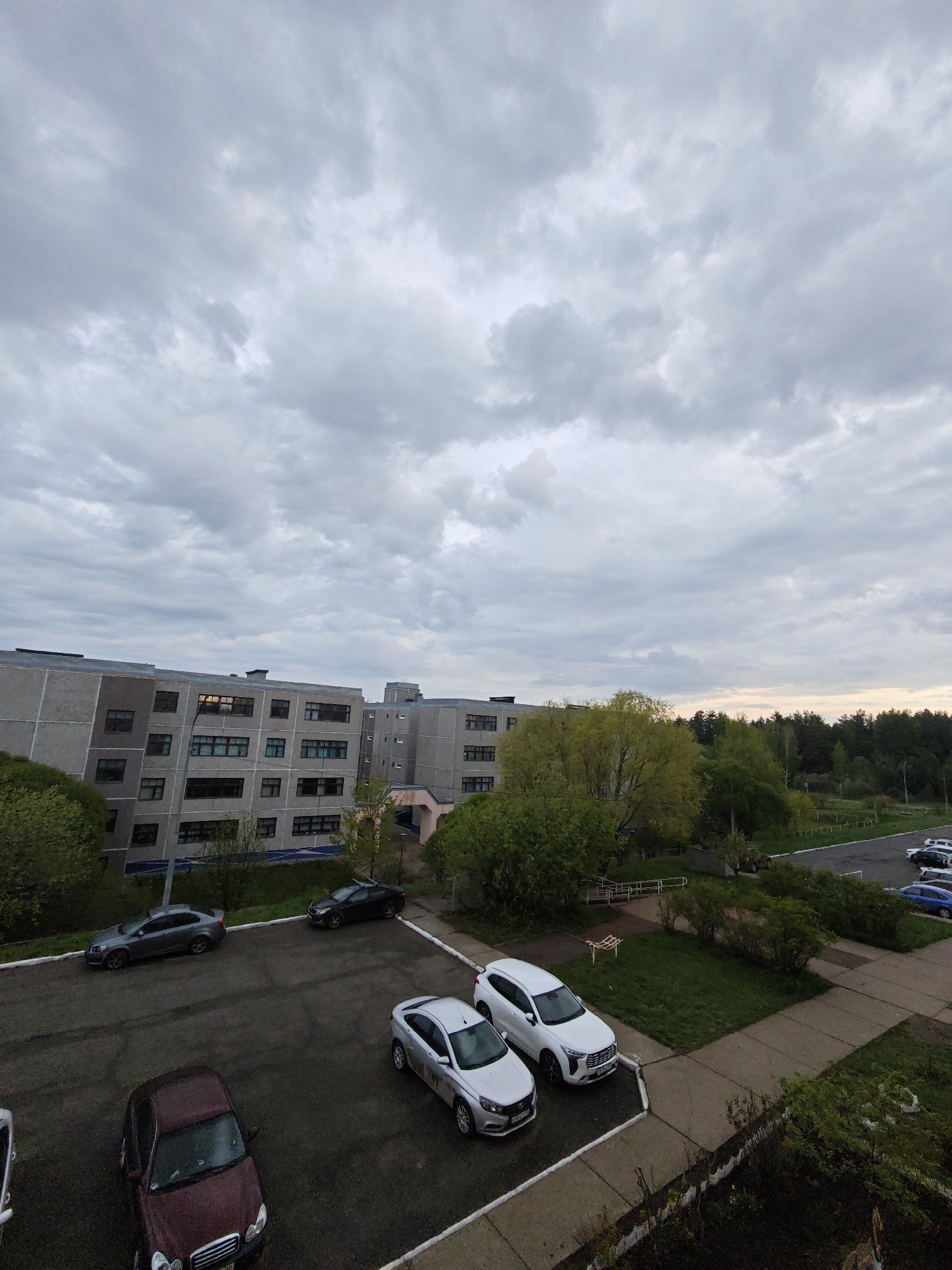 The image depicts an overcast outdoor scene in Markovskii, Russia. The foreground contains a parking lot with several parked cars, including a maroon sedan, two silver sedans, a white SUV, a black sedan, and a grey sedan. A multistory concrete building is visible in the midground, and a line of trees borders the edge of the frame on the right side. The sky is mostly cloudy.