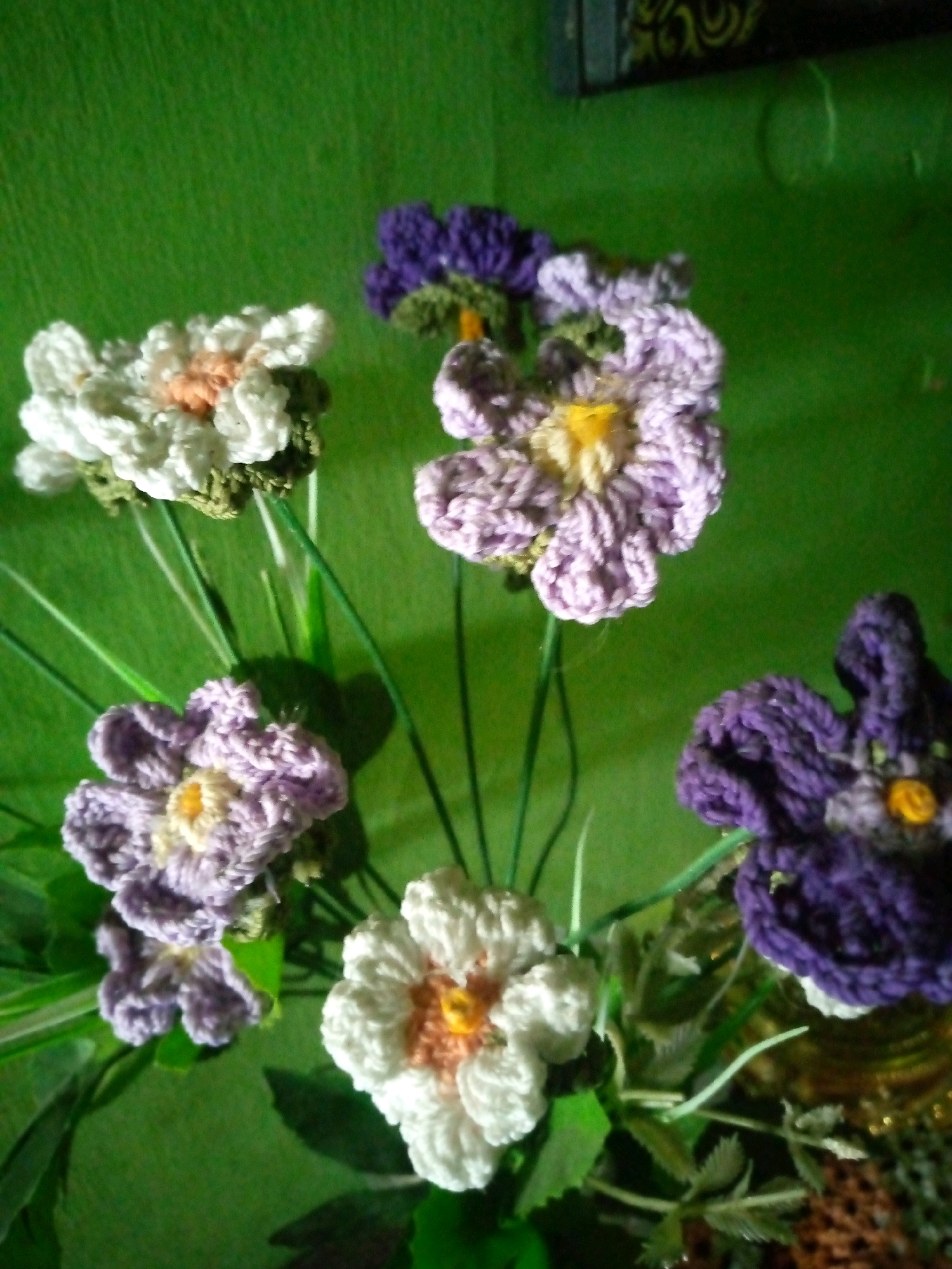In the city of Sumberwono, Indonesia, a close-up shot captures a bouquet of crocheted flowers against a vibrant green backdrop. The flowers are meticulously crafted, with delicate petals in shades of white, lavender, and deep purple. Some flowers feature yellow or peach centers, adding pops of color and depth to the arrangement. The background is a textured green surface, possibly a wall or fabric, which contrasts with the intricate textures of the crocheted flowers. The lighting casts subtle shadows, highlighting the three-dimensional nature of the handcrafted blooms and creating a soft, almost dreamlike atmosphere.