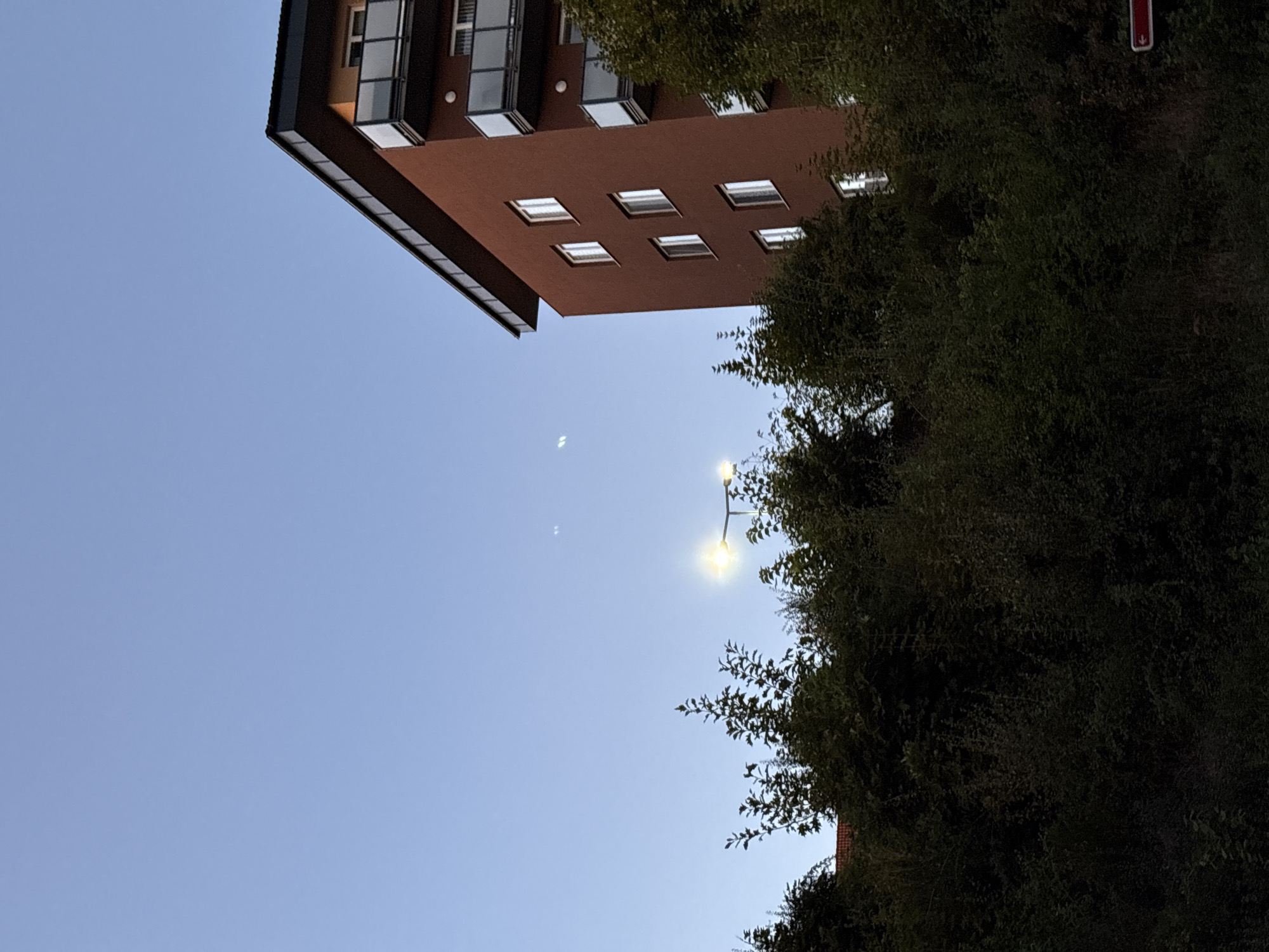 A tall, reddish-brown apartment building is situated to the right of the frame, with a dark roofline and multiple floors. Each floor has windows, some with balconies featuring glass railings. Trees and dense foliage are prominent in the foreground and to the left of the building, obscuring much of the lower portion of the structure and the ground. A street lamp with a bright light is visible above the trees, and two faint, illuminated objects are present in the sky above and to the left of the lamp. The sky is clear and light blue, suggesting it is either early morning or late evening. A red sign with a white arrow pointing left is partially visible in the lower right corner. The scene is an exterior view of a residential area with a clear sky and some artificial lighting.