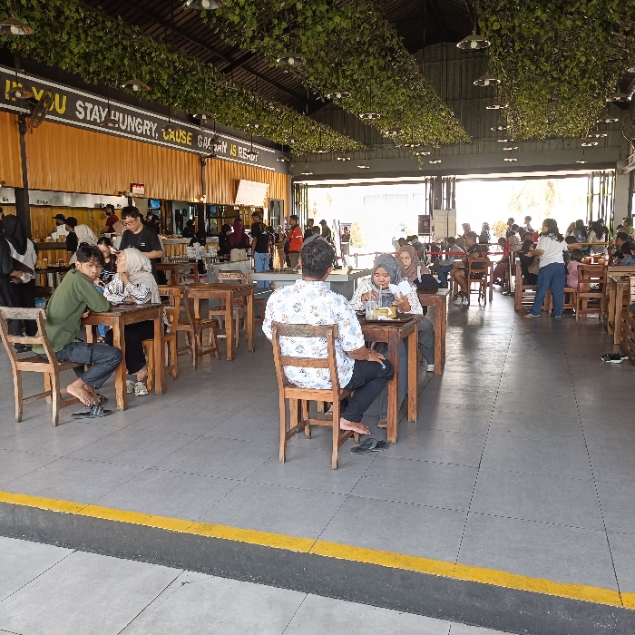 The scene is an outdoor restaurant in Kecamatan Ngaliyan, Indonesia, located at latitude -7.002 and longitude 110.342. It is partially covered with a leafy canopy. Several tables are set up with people sitting and eating. Many people are standing and talking near the service counter. There is a large yellow sign with the phrase "If you stay hungry, cause hungry is rest". The floor is tiled, and there are yellow lines on the floor. Several people are standing around and others are seated in the shade, and most of the people are facing each other, likely interacting. Some people are wearing hats, shirts, and trousers while others are wearing traditional clothing. There are several wooden tables and chairs set up. A few people are carrying small bags. The weather appears to be clear, and the light is bright.