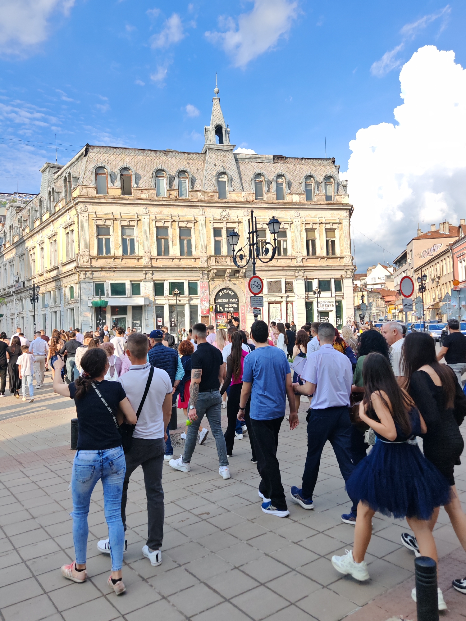 The image shows a bustling scene in Ruse, Bulgaria. A large crowd of people is gathered in a paved square. The building in the background is the Dohodno Zdanie, a historic architectural landmark in the city. The building is a light color, with ornate detailing, a distinctive spire, and multiple windows. A tall, decorative lamppost is also visible. The people in the foreground are of varying ages and are dressed in casual clothing. Some are walking, while others appear to be standing or interacting with one another. The sky is partially cloudy. Signs in Bulgarian are visible.