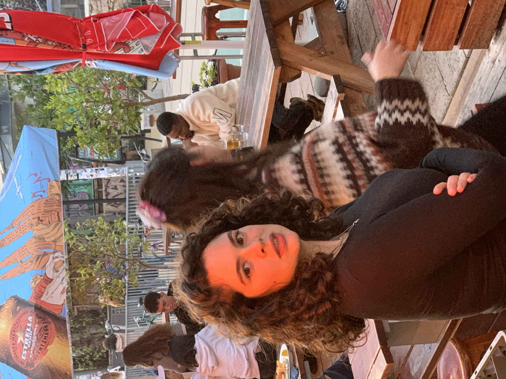 The image captures an outdoor seating area at Fuentzman Bar, located on J. L. Gordon Street, Tel-Aviv, Israel, during what appears to be late afternoon or early evening.
In the immediate foreground, a young adult female with dark, curly hair and a nose piercing looks directly at the camera, her face partially illuminated by an orange light source. She is wearing a dark, long-sleeved top with her arms crossed. To her right, another female, seen from behind, wears a patterned brown, white, and red striped sweater, with a pink hair clip visible in her dark hair. Her right arm is extended towards a wooden box on the table.
Multiple wooden picnic-style tables with attached benches are arranged across the scene. At a table further back and to the right, a dark-skinned male with a short beard and haircut is seated, wearing a cream-colored top with a dark "M" logo on the sleeve. He is seen with his right hand near his mouth, looking down or to his left, with a glass of beer and another clear glass on his table. Other patrons are visible in the background and mid-ground, including a male looking downwards at a table to the left and a female in a white hoodie with her back to the camera. On a table to the left of the foreground subject, a white plate containing what appears to be fried food with a lemon wedge and a pair of sunglasses is visible.
Overhead, a large blue open umbrella features "ESTRELLA DAMM BARCELONA" branding along with the name "FUENTZMAN" and an architectural illustration. Another blue umbrella with musical notes is partially visible, while a large red and blue umbrella with Hebrew text is folded and tied on the right side of the frame. Small potted trees and plants are scattered throughout the seating area, and a grey metal fence separates the bar from a paved walkway where several bicycles are parked. A wall adorned with graffiti and posters can be seen in the background to the left. The ground is paved with light-colored interlocking blocks.