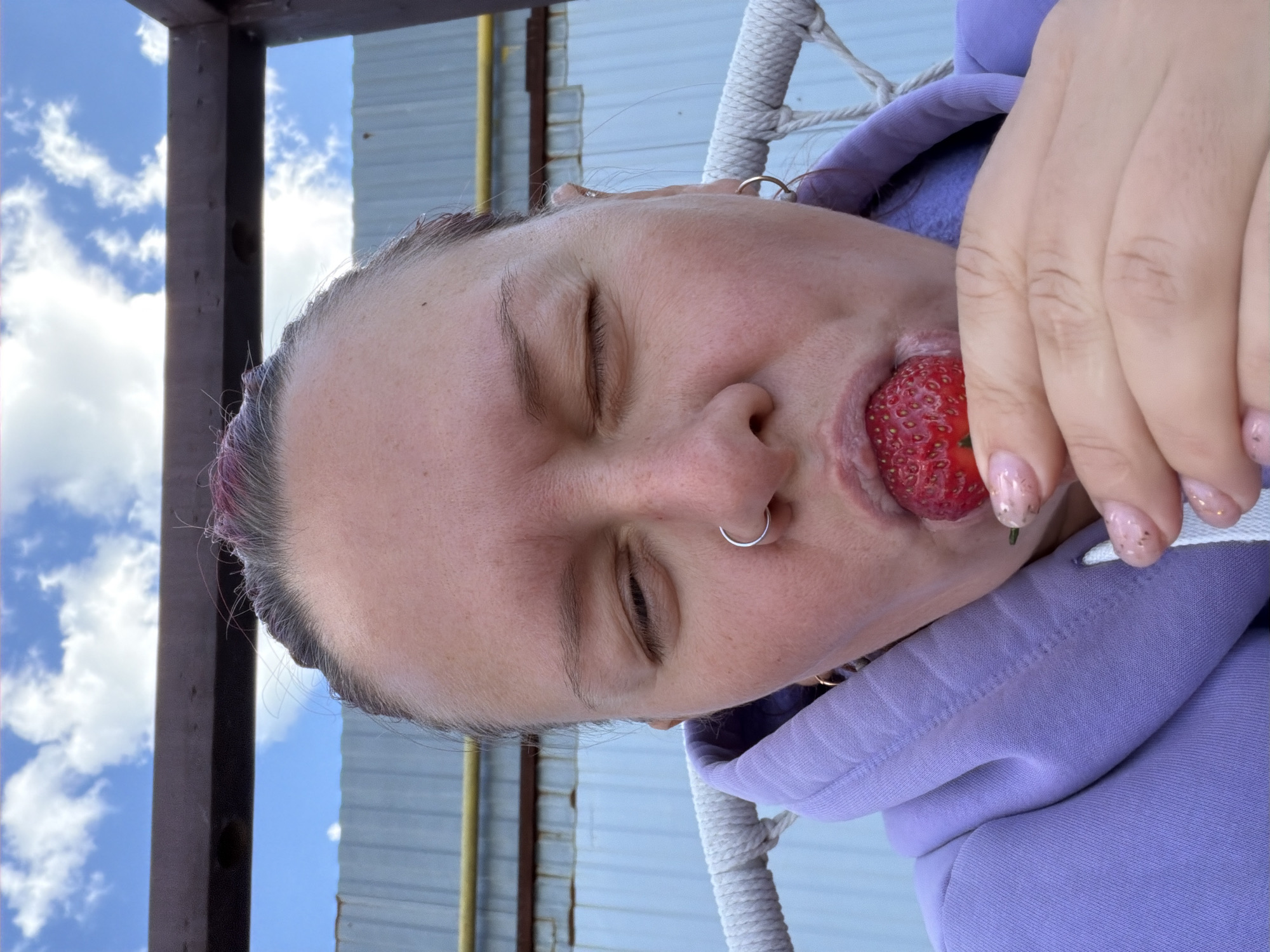 A person with light skin and closed eyes is holding a strawberry in their mouth. They have a nose ring and are wearing a purple hoodie. The background shows a metal structure, a light blue sky, and white clouds. The location is Chaikovsky, Russia.