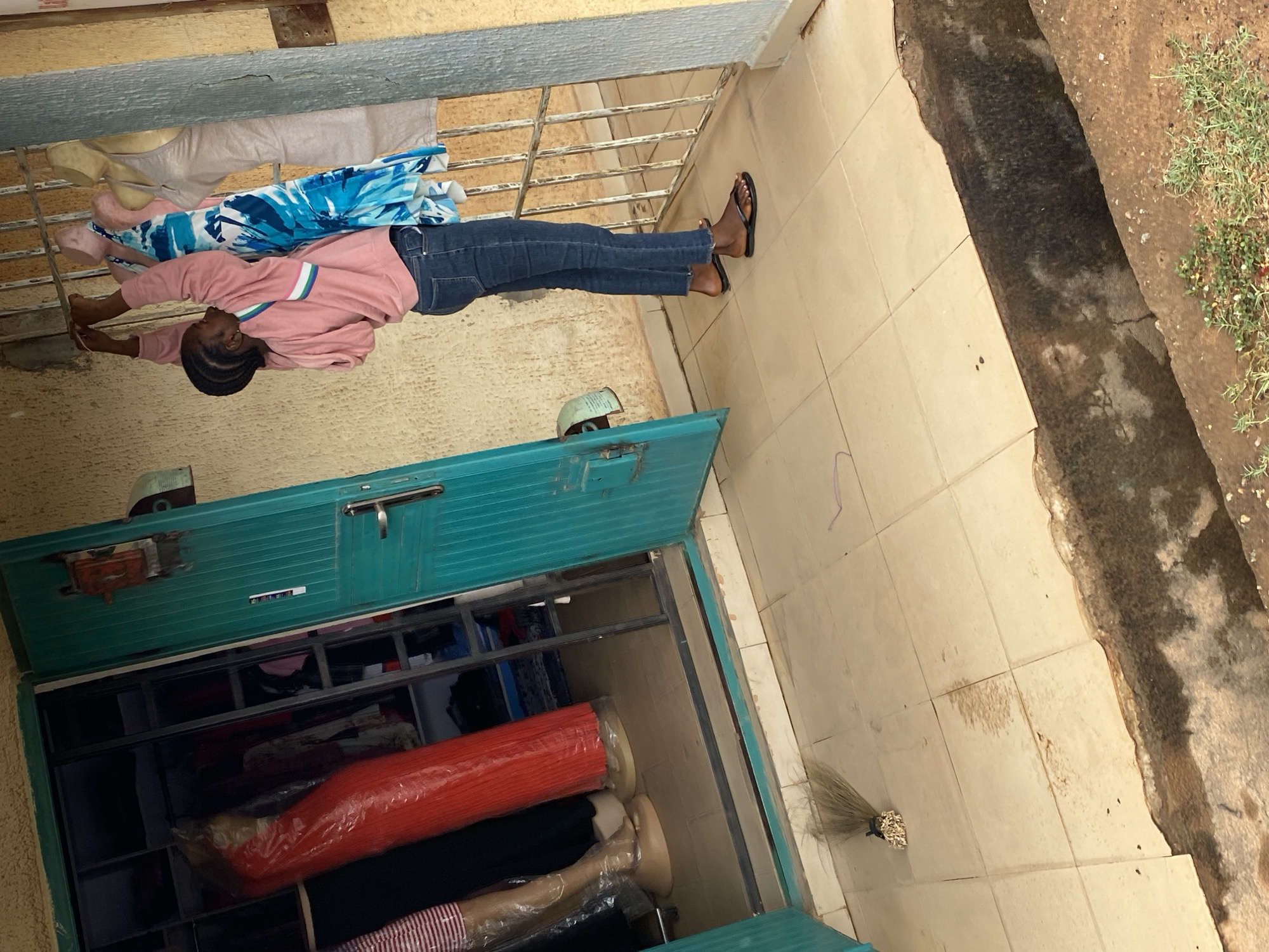 A woman is standing outside a building in Narayi, Nigeria. She wears a pink long-sleeved shirt, blue jeans, and flip-flops. She stands in front of an open, turquoise-colored storage unit filled with clothing items, including red, black, and striped textiles. The unit is metal-framed and has a metal door with a handle. Next to the unit, on tiled flooring, there is a broom. Outside, there is a concrete drain with some vegetation. The building walls and metal bars are visible in the background.