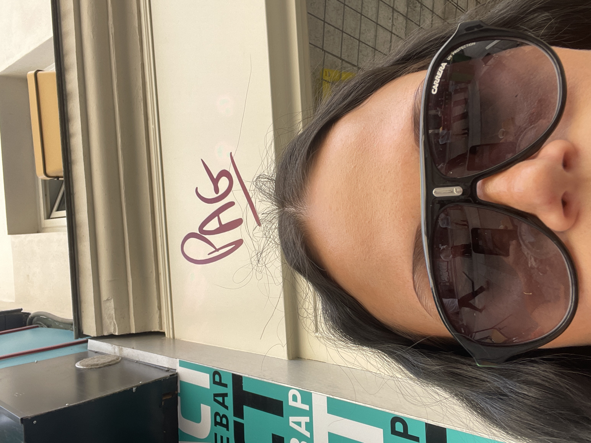 A close-up, tilted shot of a person wearing Carrera sunglasses, obscuring much of their face, taken outdoors next to a wall covered in graffiti. The graffiti includes the word "PAG" in purple writing. Below the graffiti is a green and white advertisement with "BAP" partially visible. The environment includes what appears to be a Parisian building's exterior with stone molding. Reflected in the sunglasses are images suggesting an urban setting, possibly including a person and street furniture.