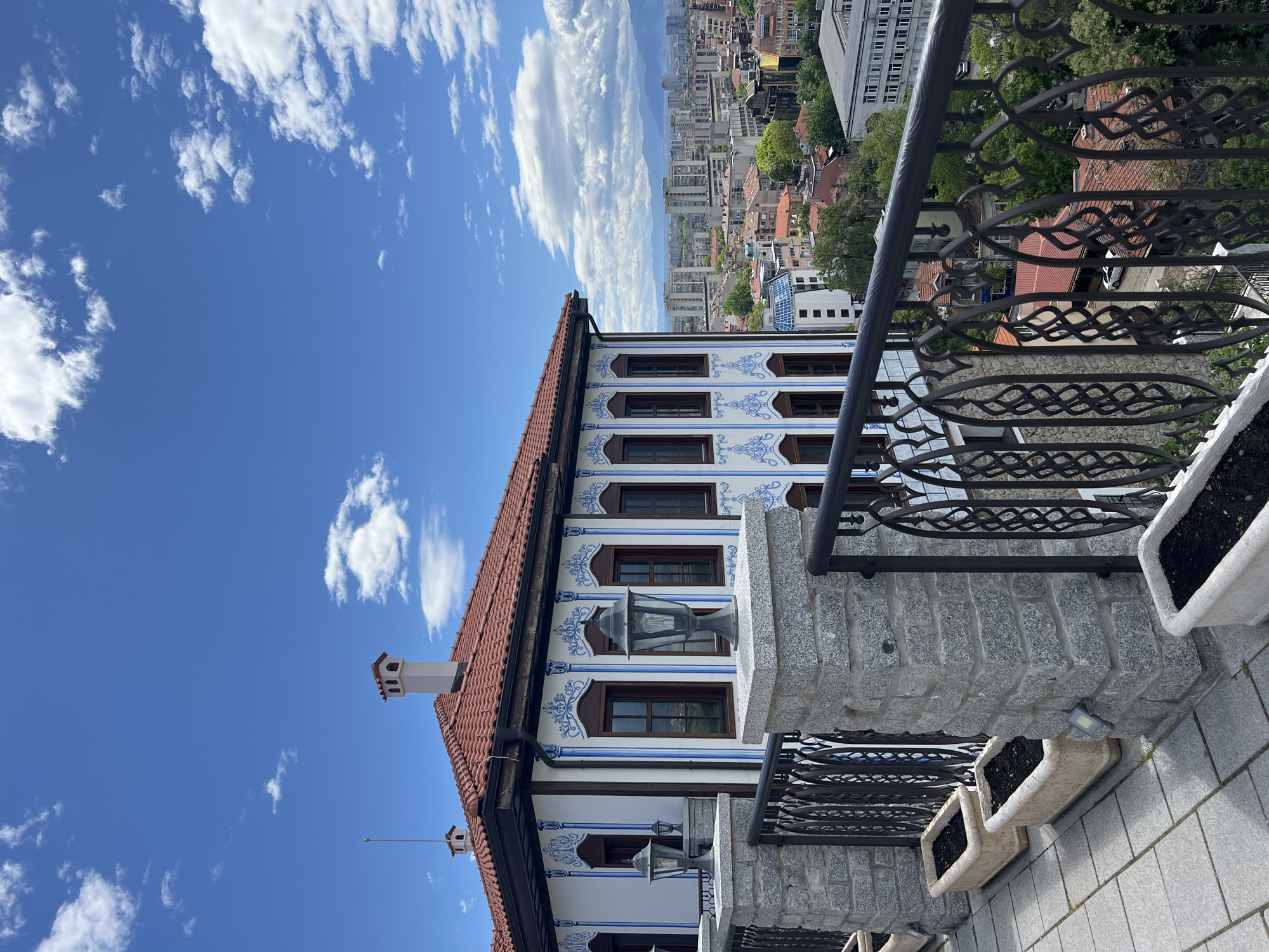 The image depicts the exterior of the Georgiadi House in Plovdiv, Bulgaria, a notable example of Bulgarian National Revival architecture. The building features a distinctive façade with white walls adorned with blue floral patterns around the windows. A red-tiled roof is visible, with a chimney at one end. A wrought iron balcony with a stone base is in the foreground. Beyond the house, a cityscape stretches out, with buildings of varying heights and architectural styles. The sky is blue with scattered white clouds. The photo is taken from a high vantage point, looking slightly downward.