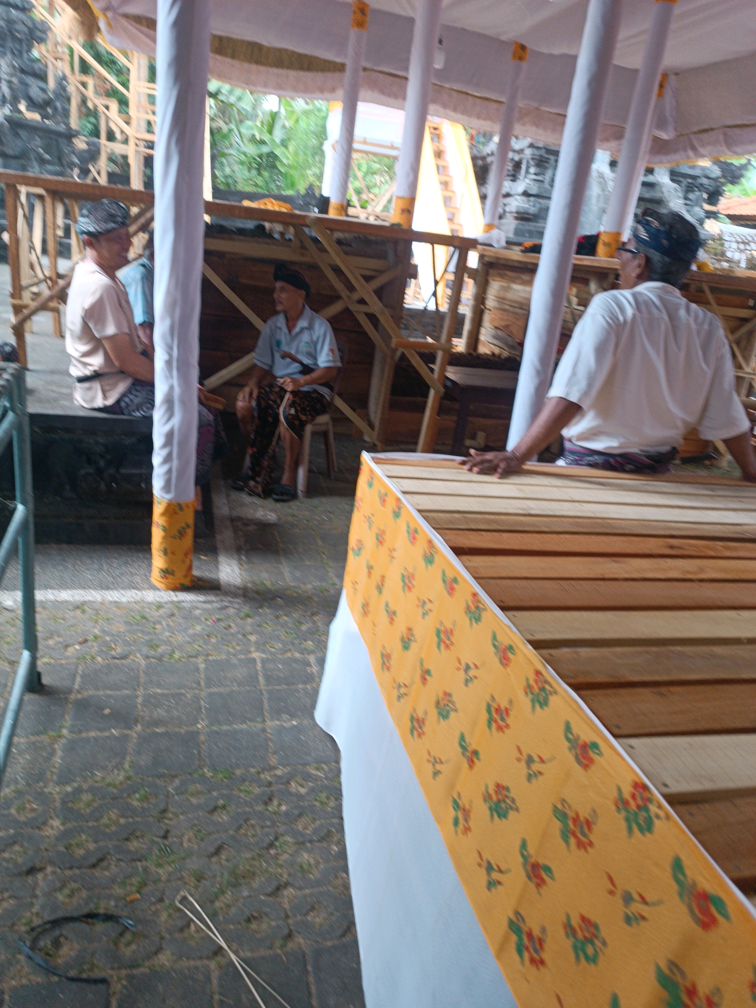 The image depicts an outdoor scene possibly in Selemadeg, Indonesia, under a temporary canopy with white fabric and yellow accents. In the foreground, a person wearing a white shirt and a patterned waist sash is leaning over a wooden structure, their left hand resting on the planks. The wooden structure is covered with a yellow fabric adorned with a floral print in red and green. To the left, a pillar wrapped in yellow fabric with a similar floral pattern stands. Further back, two men are seated. One, wearing a light-colored shirt and a dark patterned sarong, is looking to the left. The other, in a light blue shirt and a dark patterned sarong, is seated and holding a long, thin object, possibly a stick or a rod. Behind them, wooden structures and foliage are visible. The ground is paved with grey interlocking bricks.