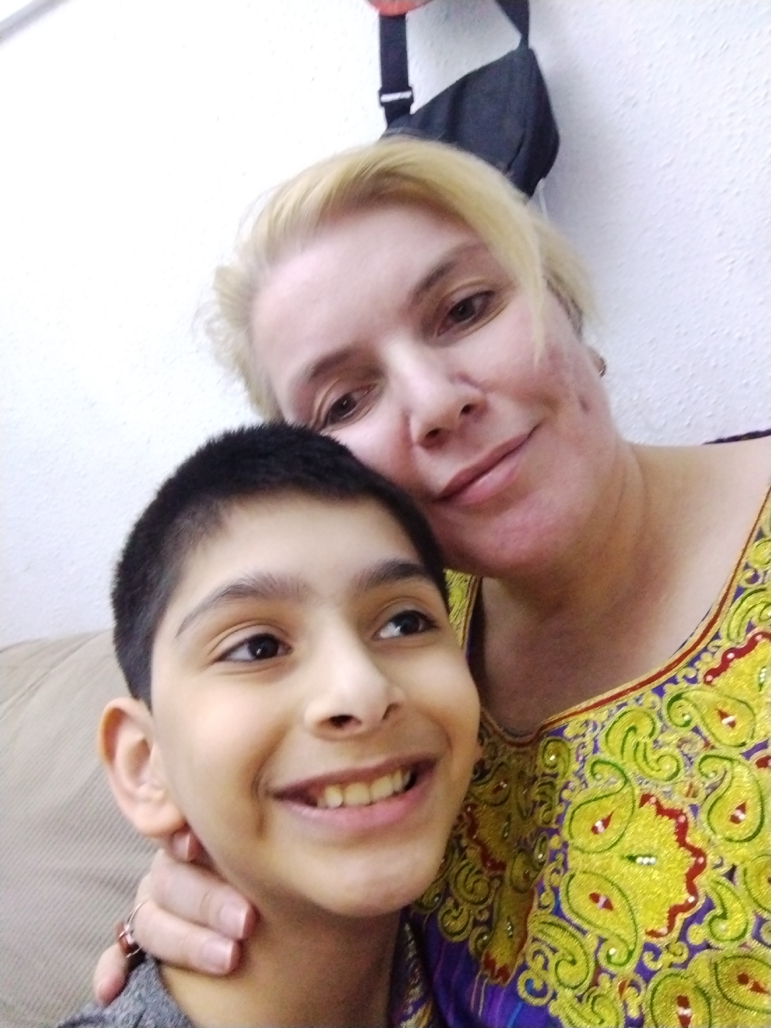 In the image, a woman with blonde hair and a boy with dark hair are posing for a selfie. The woman is to the right, with her head tilted and her arm around the boy. The boy is on the left, smiling directly at the camera. They are both wearing casual clothing. The background appears to be an indoor setting with a textured wall. A black bag is visible in the upper right corner. The overall lighting suggests the photo was taken indoors with artificial light. There is no visible indication of the location being Al Mas'oudi, United Arab Emirates.