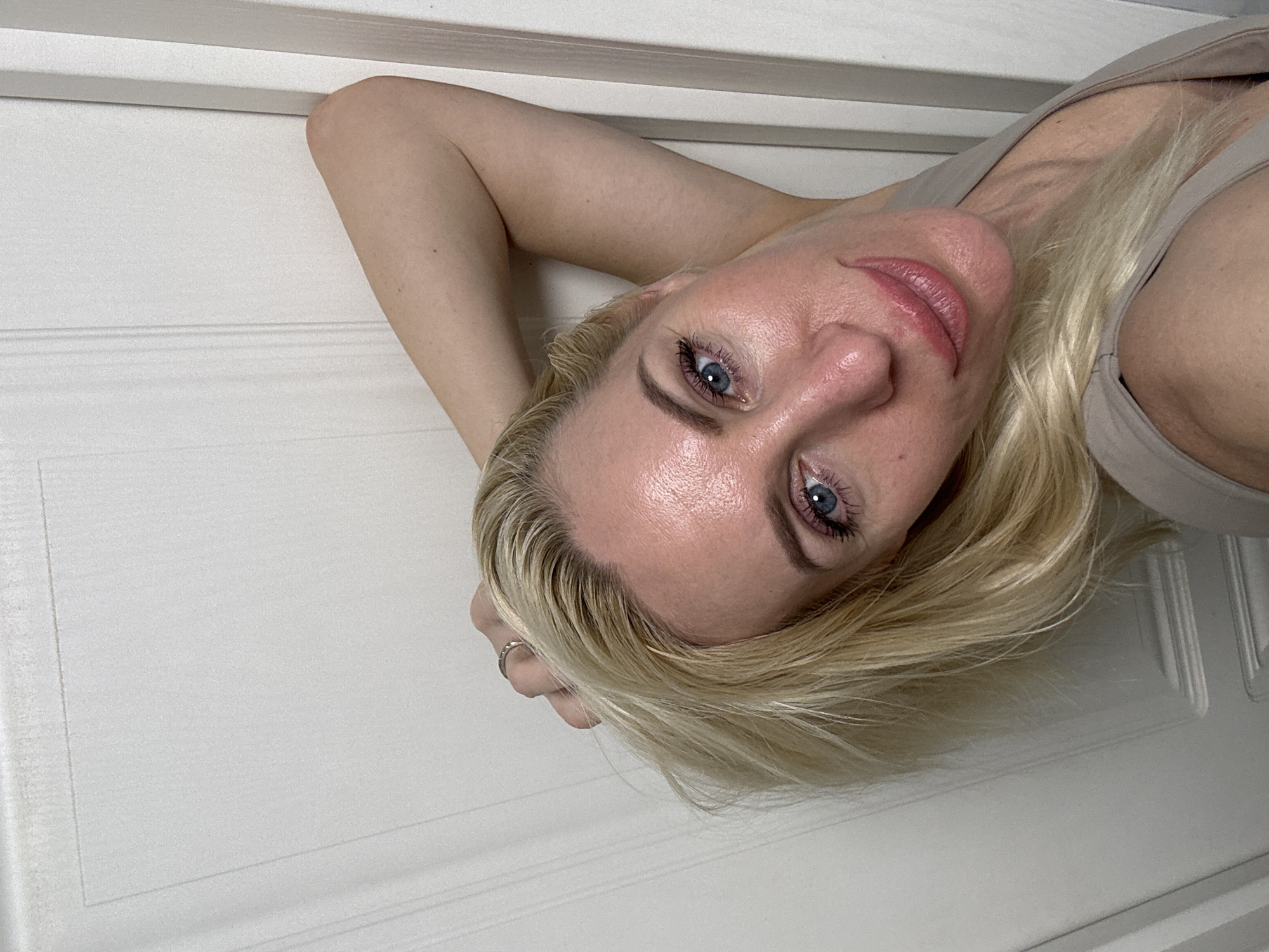 A blonde woman is captured in a selfie, leaning her head against a white doorframe. Her right arm is raised and bent, with her hand resting on the back of her head, revealing a ring on her index finger. The woman's head is tilted to her left, and she is looking directly at the camera. Her makeup is visible, including mascara and a pink lipstick. The background is a plain white door with vertical paneling and a simple frame. The lighting suggests an indoor setting, possibly a home. The image is a close-up, focusing on the woman's face and upper body.