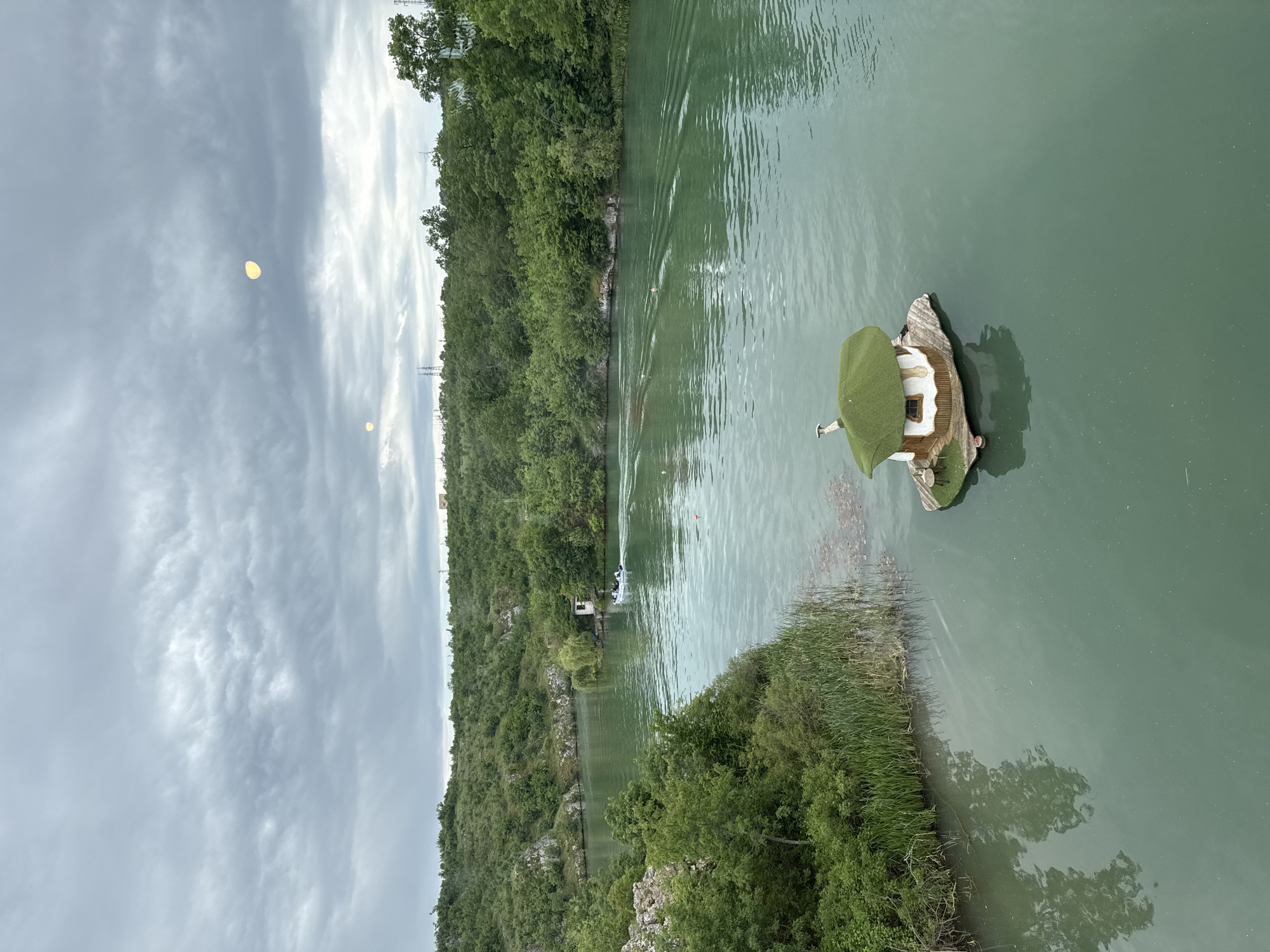 A vertically-oriented photograph captures a scene from Lukovit, Bulgaria, showcasing a river landscape under a cloudy sky. The river, a prominent feature, is flanked by dense greenery on both sides, suggesting a natural setting. A small, whimsical house structure sits on a floating platform in the water. A group of people in a small watercraft are visible on the river. Utility poles can be seen behind the trees.