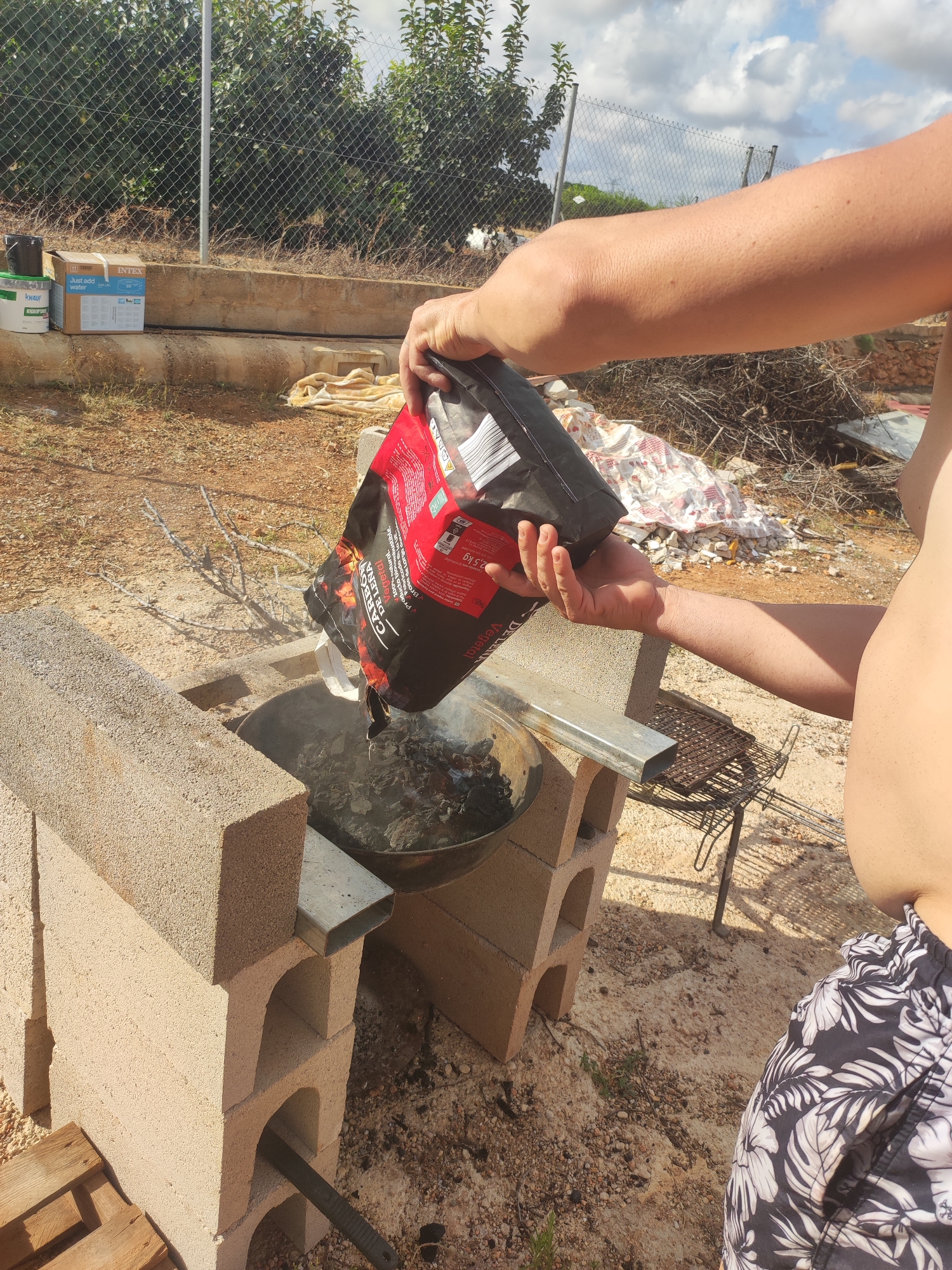 The image shows a person's arm and torso from the chest up, wearing patterned shorts, holding a bag of charcoal over a barbecue pit. The barbecue pit is constructed from concrete blocks. Ash and embers are visible in the pit. In the background, there is a chain-link fence and some vegetation. Several cardboard boxes are stacked on a concrete ledge to the left of the frame, and a rolled-up blanket or tarp is on the ground behind the barbecue. The ground is covered in gravel and dirt. The scene appears to be outdoors in a dry, sunny climate. There are no specific landmarks or details in the image that would definitively place it in Alcàsser, Spain, or any other specific city.