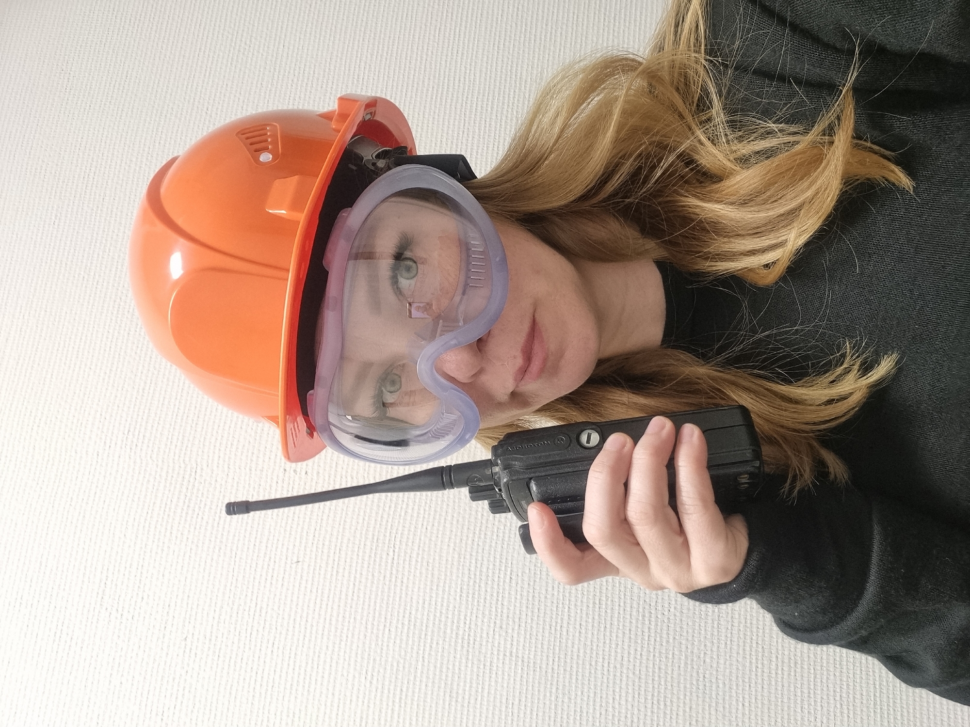 A woman is wearing an orange hard hat and safety goggles. She is holding a black Motorola walkie-talkie. The background is a textured white surface. The location is not discernible from the image.