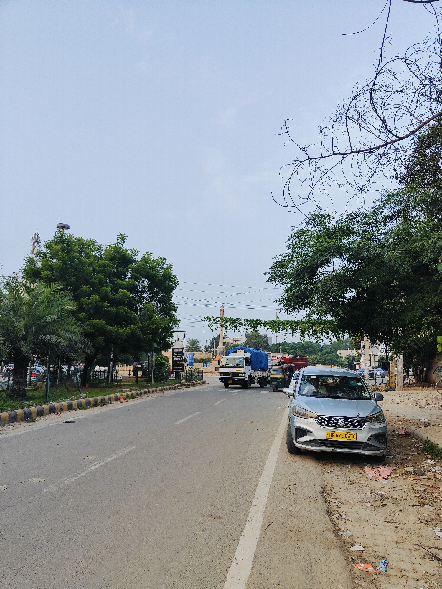 The image depicts a street scene in Gurgaon, India. In the foreground, a silver car with a yellow license plate reading "HR 67E 8450" is parked on the side of the road. The road itself is paved and has a white dividing line. To the left of the road, there is a sidewalk with a yellow and black striped curb. Beyond the curb, there are trees and a fence. In the background, several trucks and an auto-rickshaw are driving on the road. There are also buildings and telecommunication towers visible in the distance, as well as more trees and vegetation. The sky is overcast with a pale blue color. The scene appears to be a typical urban environment with traffic and roadside amenities.