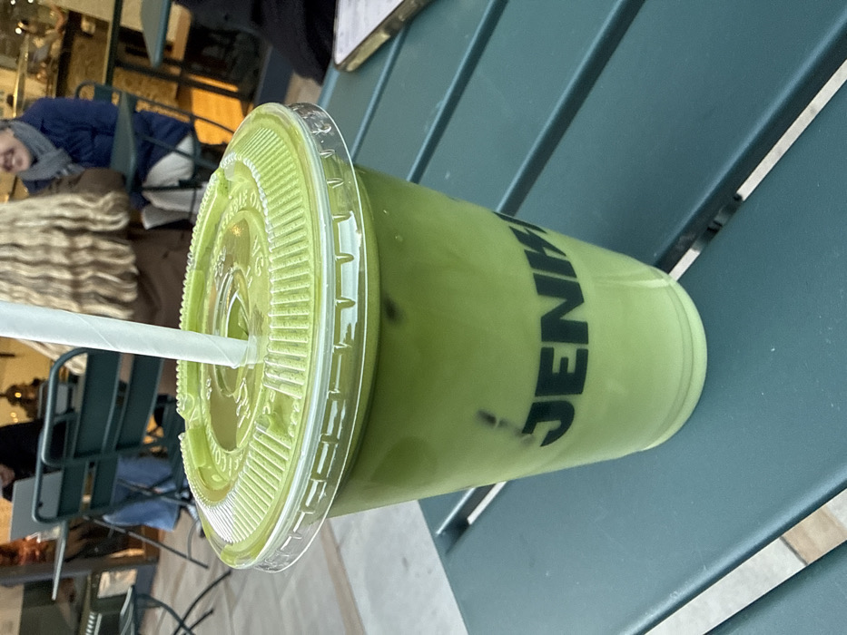 The image displays an iced green beverage in a clear plastic cup, branded "JENKI" in black capital letters, placed on a dark teal slatted outdoor table. The drink, appearing as a matcha latte, contains dark ice cubes and is topped with a light green plastic lid and a white paper straw. In the blurred background, several individuals are seated at similar dark green metal bistro tables and chairs. One person with long, wavy light brown hair is visible from behind. To their right, another individual wearing a blue jacket and a grey scarf is partially visible, smiling. Further left, another person is seen interacting with a laptop. The scene depicts an outdoor cafe setting, identified as a JENKI matcha bar in London, United Kingdom.