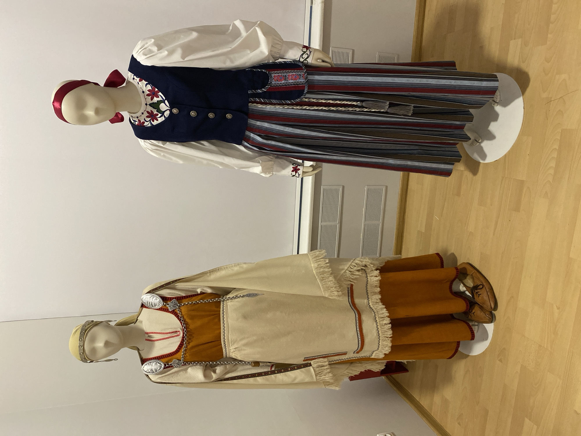 The image displays two mannequins dressed in traditional Karelian folk costumes, positioned in a museum exhibit. The mannequin on the left wears a beige tunic-like dress over an orange skirt, adorned with decorative silver brooches and chain. It also has a fringed shawl. The mannequin on the right is dressed in a white blouse with voluminous sleeves, a dark blue vest embroidered with floral motifs, and a long, striped skirt. A red ribbon is tied around its head. The background is a plain white wall, and the floor is made of light-colored wooden planks. This exhibit is located at the National Museum of the Republic of Karelia in Petrozavodsk, Russia.