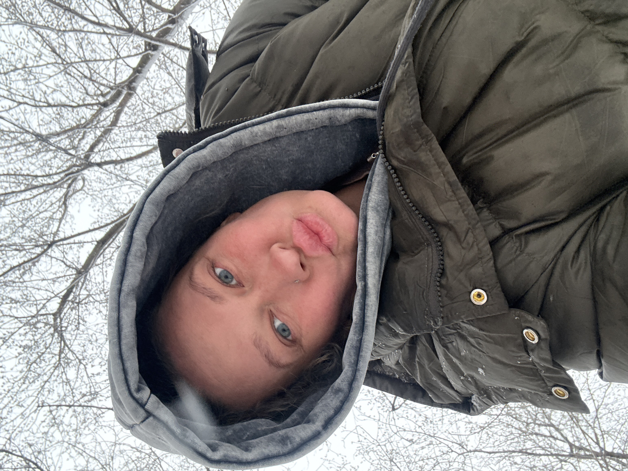 The media file captures a close-up, low-angle selfie of a person, likely a young adult, outdoors in a cold, wintry setting, characteristic of Perm, Russia. The individual's head is tilted or rotated, making them appear upside down relative to the background, as they look directly at the camera.
The subject has light skin, blue eyes, and light brown or blonde hair visible around their face. They are wearing a dark olive green puffer jacket with a grey hooded sweatshirt underneath. The jacket features visible zippers and metallic snaps. Their lips are slightly puckered in a soft pout, and a small silver piercing is present on their nose.
The background is filled with a dense pattern of bare tree branches, heavily coated in frost or snow, indicating cold, icy weather. The sky appears overcast and bright. The heavy winter clothing worn by the person further emphasizes the cold conditions. No text is visible in the image.