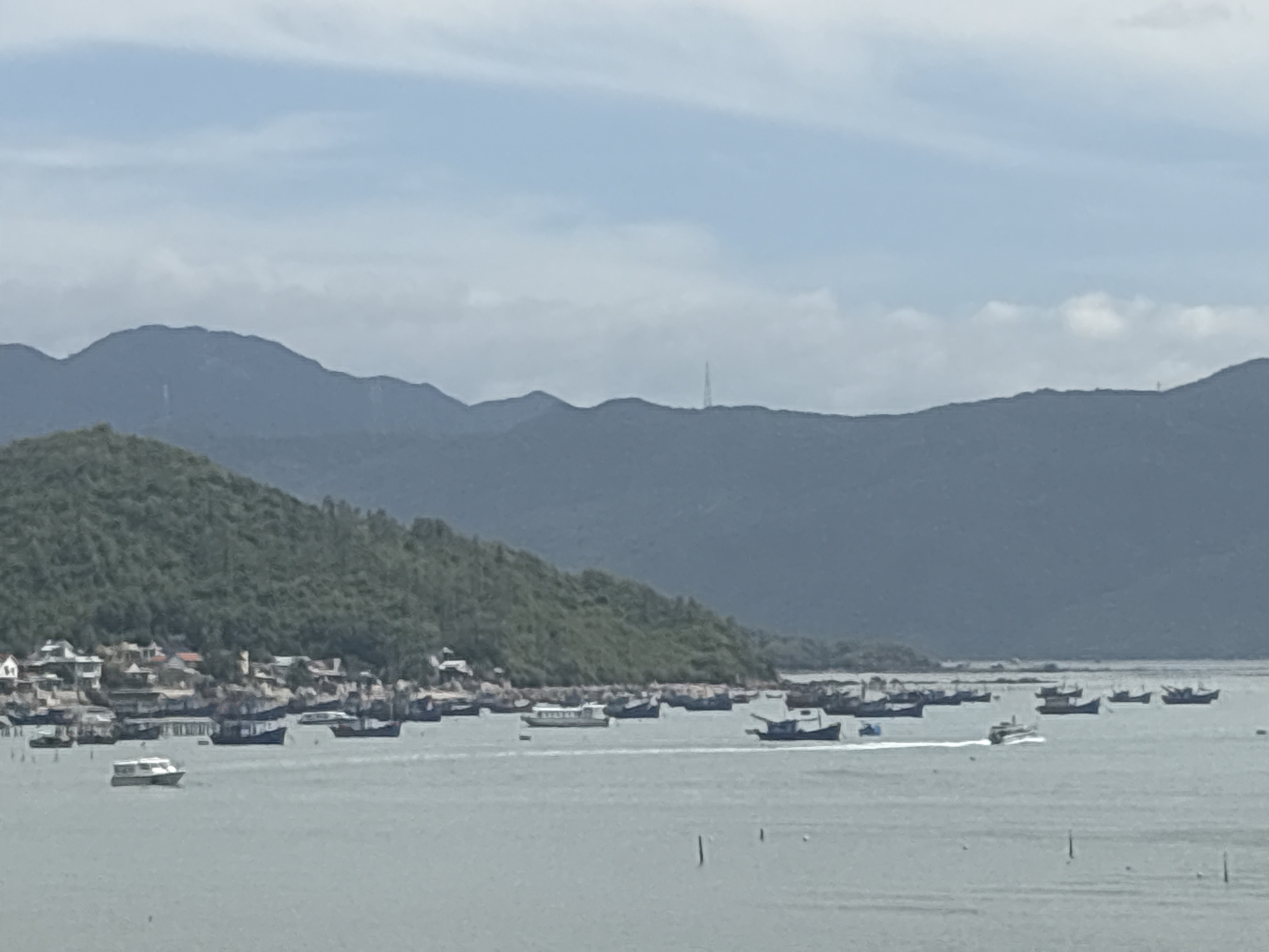 This is a wide, panoramic view of a coastal scene in Bắc Nha Trang, Vietnam, on a cloudy day. The foreground is dominated by a calm, grayish-blue body of water, dotted with numerous boats of varying sizes. Many are small fishing vessels, some appearing to be docked or anchored near the shore. A few larger boats, possibly ferries or passenger boats, are also visible, with one leaving a white wake as it moves across the water.
In the midground, a lush, green, tree-covered hillside slopes down towards the water. Along the base of the hill, a densely packed village or town is nestled, with a collection of buildings, many with red or brown roofs, situated very close to the water's edge. Some structures appear to be built on stilts over the water, suggesting a waterside community.
The background features a series of layered, dark blue mountains, with softer, lighter blue sky above. There are scattered white clouds, and a tall, slender transmission tower is visible on one of the distant mountain peaks. The overall impression is one of a bustling coastal community situated against a dramatic natural landscape.