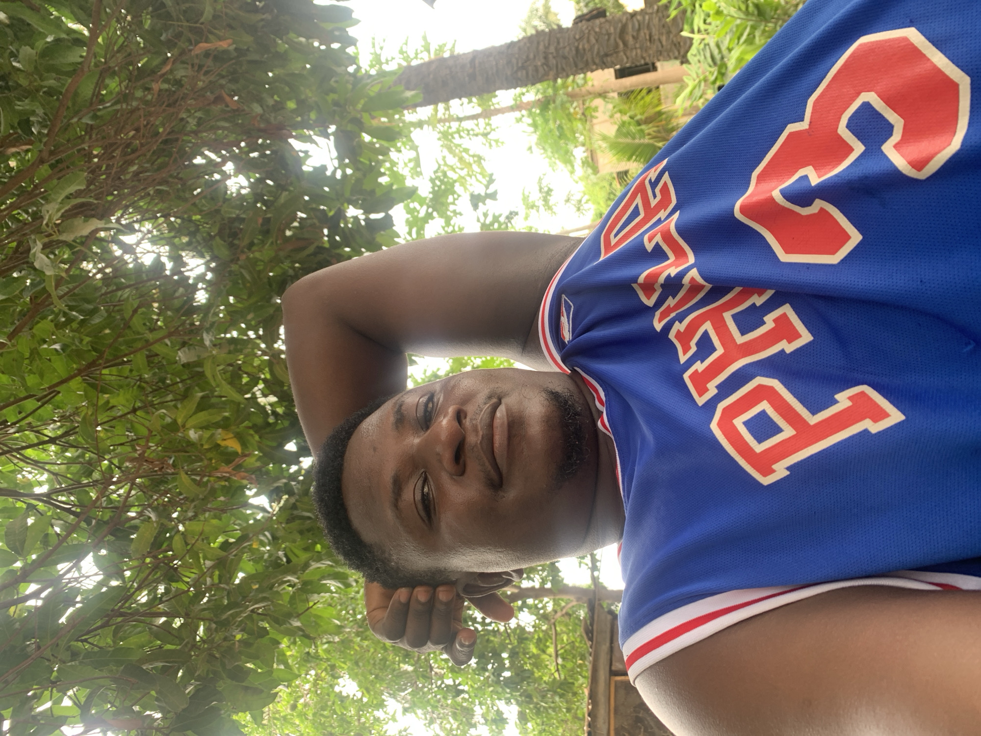 The media file features a portrait of a dark-skinned male in an outdoor setting, taken in Akwanga, Nigeria. The image is oriented sideways, showing the subject with his head tilted towards the viewer, appearing to look up from a low angle. He has short dark hair, a light beard or stubble, and a relaxed, friendly expression with a slight smile. His right arm is bent, with his hand resting near his ear.
He is wearing a blue basketball jersey with red and white striped trim. The jersey prominently displays red text and numbers: "PHILA" is visible, along with the number "3," suggesting it is a Philadelphia 76ers jersey. A partial NBA logo can also be seen on the jersey's trim.
The background is filled with lush green foliage, indicating a natural environment with trees and leaves. Bright sunlight filters through the canopy, suggesting it is daytime. Several tree trunks, including what appear to be palm trees, are visible further in the background, consistent with a tropical or subtropical location. The overall scene conveys a casual and pleasant outdoor moment.