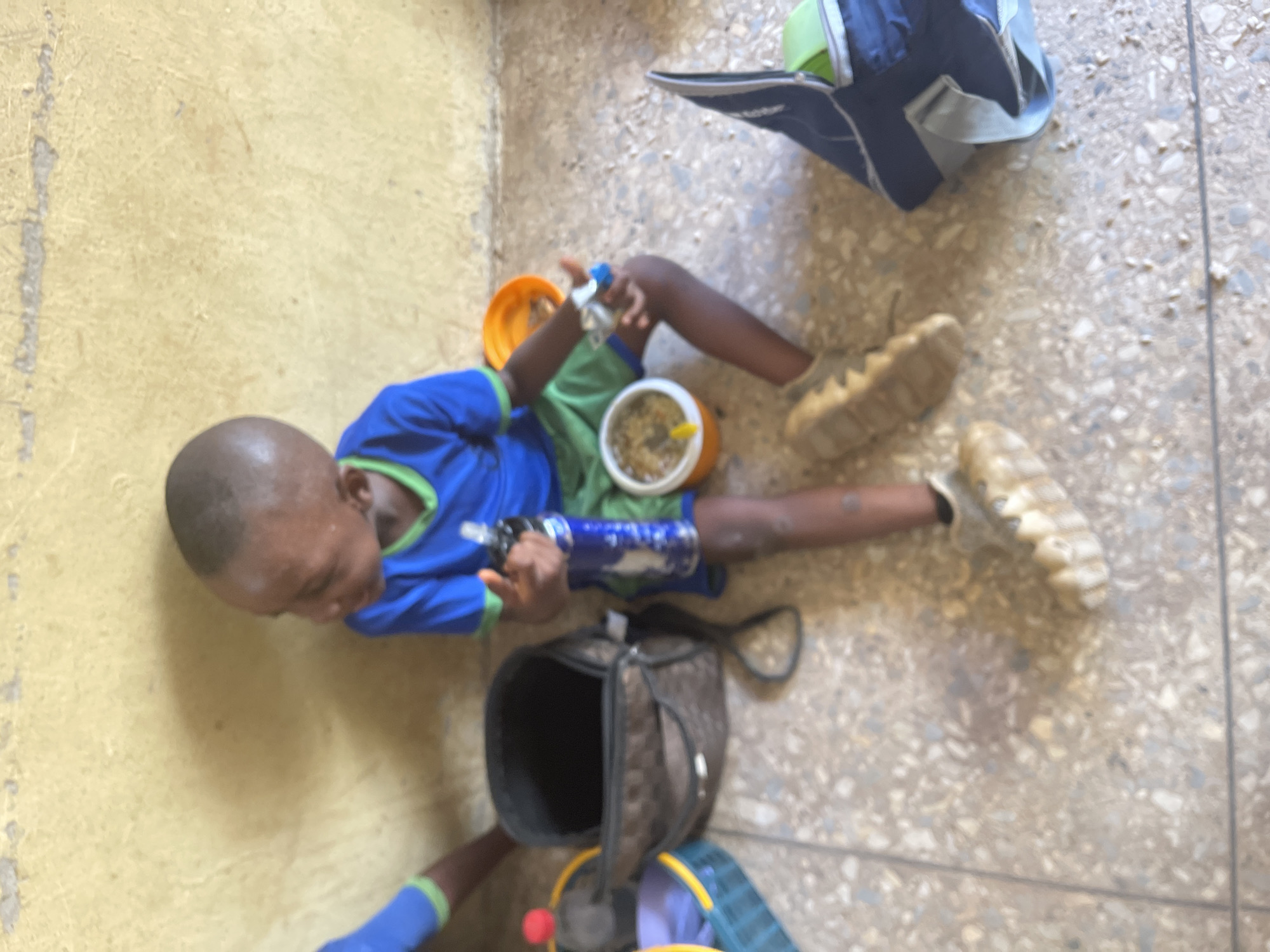 The image, captured from an overhead perspective, depicts a young person, likely a child, resting or eating on a light-colored, speckled terrazzo or concrete floor. The child has dark skin and closely cropped dark hair, wearing a blue long-sleeved shirt with green trim and green shorts. They are wearing light-colored, sturdy shoes on both feet.
The child appears to be on a break, possibly consuming a meal. In their left hand, they hold a small, clear plastic water bottle with a blue cap. Near their right leg, an open, round orange food container holds what looks like a meal of grains or rice, accompanied by a small yellow spoon or utensil. Another dark blue bottle with white markings is situated near their right arm.
Several bags are scattered on the floor around the child. A dark blue and light green backpack or sports bag is visible in the upper right quadrant. An open dark brown bag, possibly a lunch bag, lies near the child's lower body and left arm. Part of a purple backpack with yellow trim is also seen in the lower central part of the frame.
The setting suggests an indoor or covered area, with a textured, light yellow wall running along the left side. The lighting indicates it is daytime. The child's expression is neutral, and they are looking towards the upper left area of the image. No visible text is present. The scene is set in Akwanga, Nigeria.