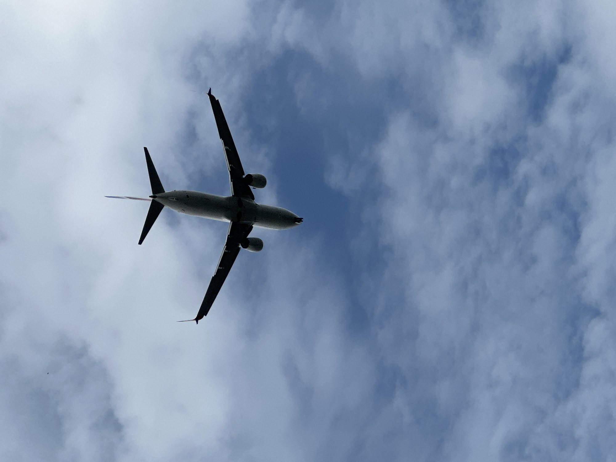 An airplane is seen from below, flying against a backdrop of a cloudy sky. The plane's underside, wings, and tail are visible. The clouds are a mix of white and gray, with patches of blue sky showing through.