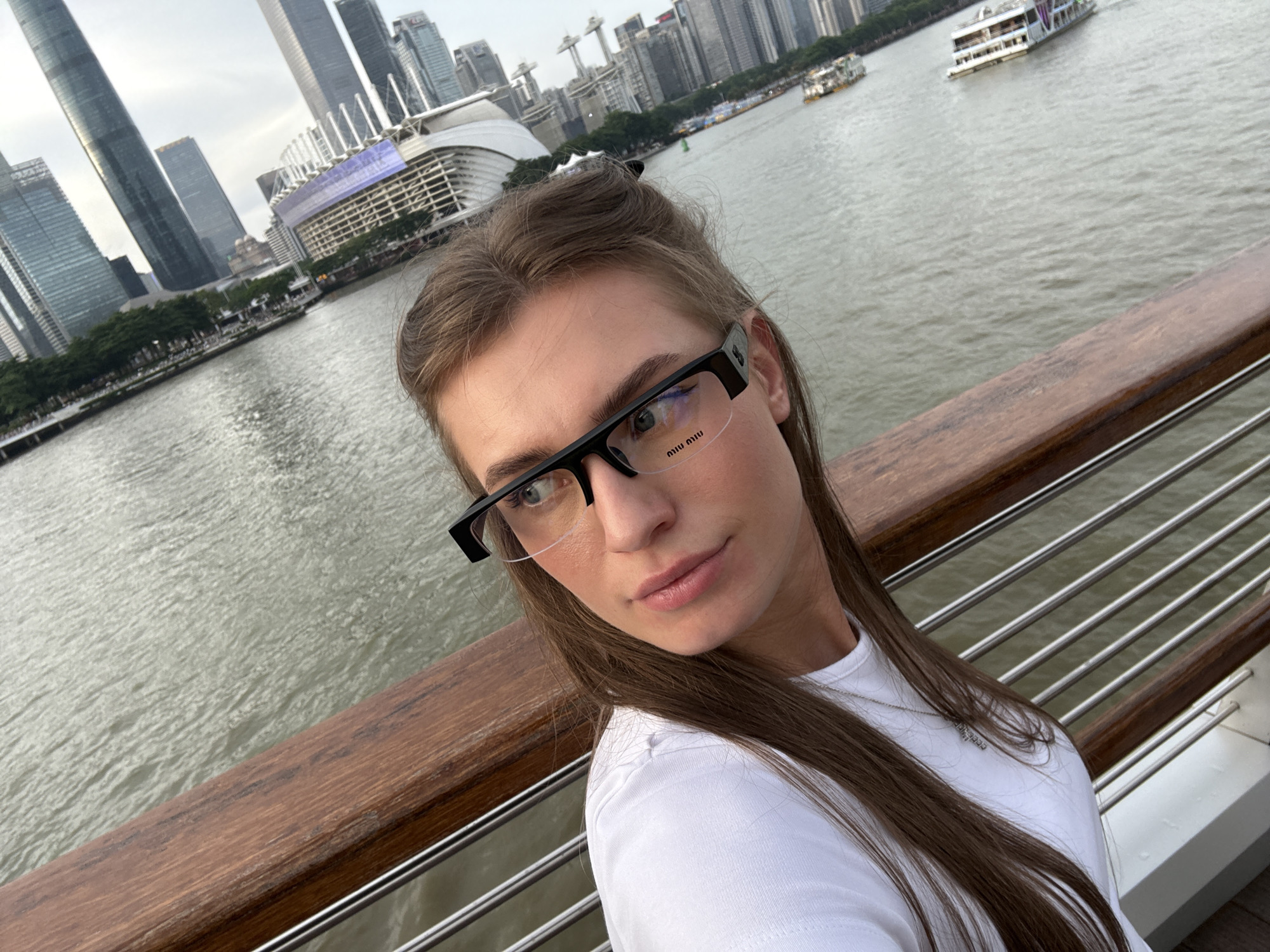 A young woman with long, brown hair and glasses takes a selfie on a wooden promenade overlooking a river in Yuexiu District, China. She is wearing a white t-shirt and a silver necklace. In the background, a cityscape with tall buildings and a large modern structure are visible across the river. Several boats, including a passenger ferry, are on the water. The sky is overcast.