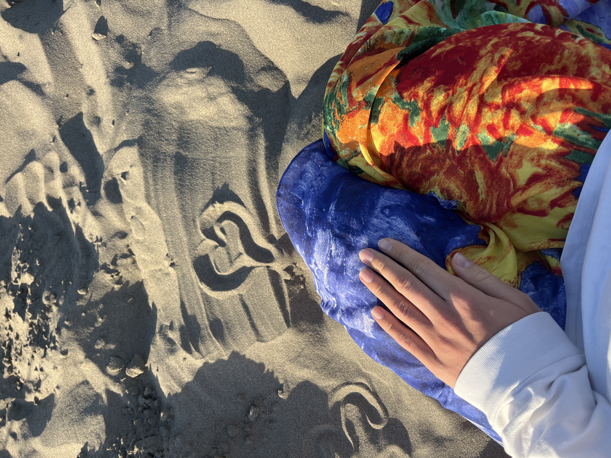 In the city of Pistula, Montenegro, a top-down, close-up shot captures a hand resting on a colorful fabric on a sandy beach. The sand, appearing light brown and textured with ripples and shadows, forms the background and foreground. A heart shape has been drawn into the sand, with the point facing towards the bottom right of the frame. The hand, belonging to a person wearing a white sweatshirt, is placed on top of two folded fabrics: one a vibrant blue with white swirls, and the other a multi-colored pattern of red, orange, yellow, and green. The scene suggests a peaceful and romantic moment on a sunny day at the beach.