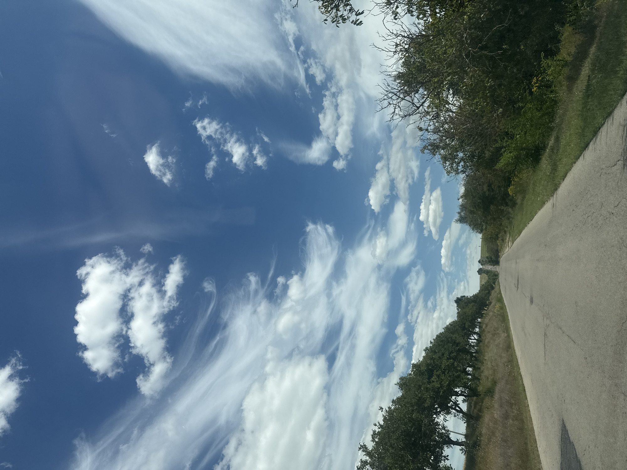 The image shows a rural road in Iurievca, Moldova, under a bright blue sky filled with wispy white clouds. The road, paved and in fair condition with some patches, stretches into the distance, curving slightly uphill. On the left side of the road, a line of mature trees with dense green foliage creates a natural border, casting shadows on the grassy embankment. The right side of the road is lined with a mix of trees and bushes, some bare-branched and others with green leaves. The landscape appears dry and rural, with fields visible in the background. The overall scene is serene and evokes a sense of journey.