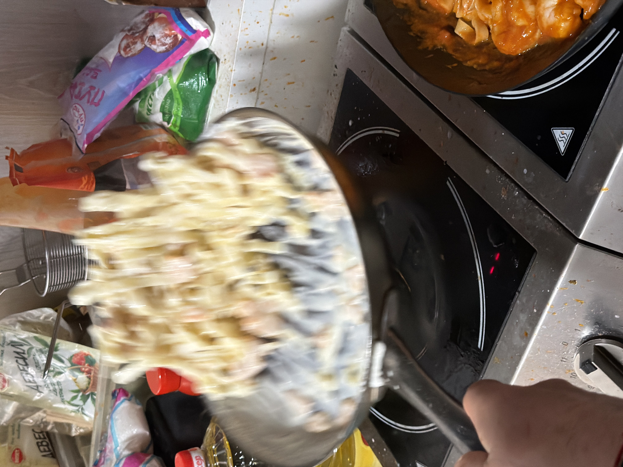 A first-person perspective photograph captures a dynamic cooking scene within a kitchen in Sofia, Bulgaria. The central action shows a person's left hand, visible in the lower-left, actively tossing a dish in a dark metal frying pan. The pan contains white or pale yellow pasta, likely fettuccine, mixed with a creamy white sauce and small, light-pink protein pieces (possibly shrimp or chicken), all in motion blur due to the tossing action.
The pan rests on a black ceramic induction stovetop. The burner beneath the active pan displays two illuminated red indicator lights, signifying it is operational. To the right, a second dark pan sits on an adjacent burner, holding pasta or gnocchi coated in a distinct orange-red sauce. Below the active pan, a stainless steel control panel for the stovetop is visible, featuring a rotary knob set near the '10' mark, with minor food splatters on its surface.
In the background, a light-colored countertop or wall exhibits reddish-brown speckles. Various kitchen items and food packages are arranged on this surface: a partially visible clear bottle with a red cap labeled "Олио" (Olio), a red spice shaker, a clear plastic bag clearly displaying the Bulgarian word "ДЕВЕСИЛ" (Lovage), a blue and pink plastic bag with "ПУРМА" visible on its surface, and a partially obscured green bag. A metal basket or strainer is situated on the far right of the background. The presence of prominent Bulgarian text on multiple food packages confirms the scene's location within Bulgaria, specifically inferred to be in a kitchen in Sofia.