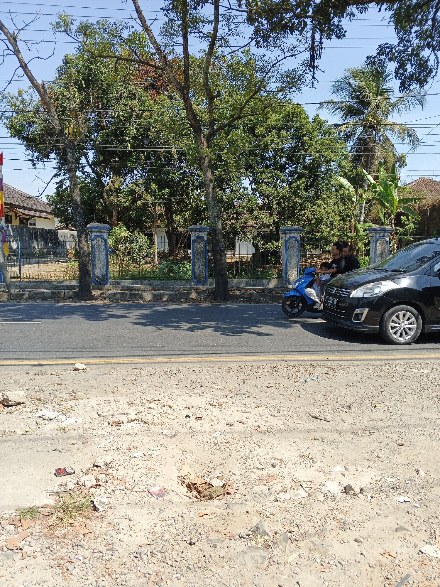 In Sukabumi, Indonesia, on a sunny day, a black car is traveling on a paved road next to a dirt shoulder. A blue scooter carrying three people is also on the road. Behind the road, a fence with decorative pillars separates the street from a property with trees and houses. The ground beside the road is covered in dirt, small rocks, and scattered debris. Overhead, power lines are strung between poles, and the sky is clear blue.