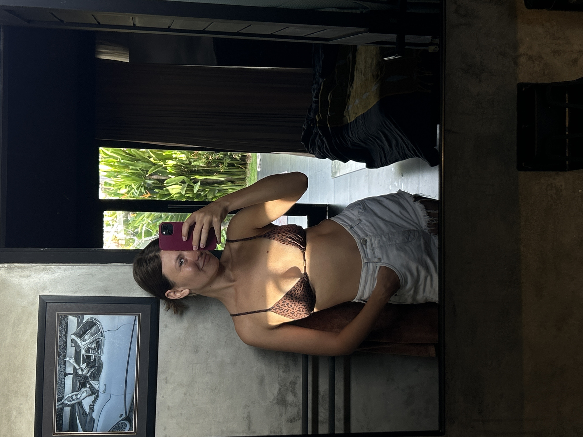 A woman in a leopard print bikini top and white denim shorts is taking a selfie in a mirror. The mirror reflects an outdoor scene with greenery, possibly in Mengwi, Indonesia. A framed black and white photograph is visible on the wall to the left of the woman. The room has a concrete wall and is dimly lit. Clothes are hanging from the top of the mirror's frame.