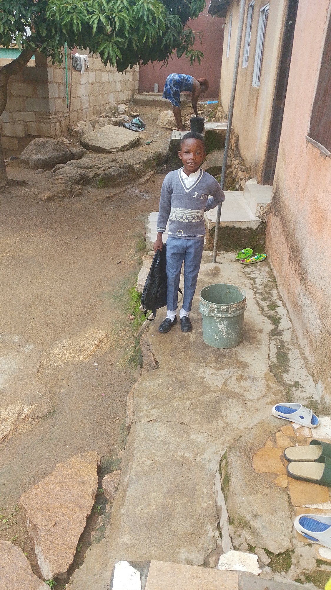 A young boy, appearing to be of school age, stands in the foreground of a dirt and concrete pathway. He is wearing a grey sweater with a V-neck and patterned trim, and blue trousers. He holds a black backpack in his left hand. To his right, a green bucket sits on the concrete. Further to the right, a pair of bright green flip-flops rests on the ground. In the background, a woman in a blue and white patterned dress is bending over near a bucket. A concrete building with windows is visible behind her. A tree with lush green leaves is to the left of the frame, casting shadows. The environment appears to be an outdoor residential area, possibly a courtyard or alleyway, with some uneven terrain and large rocks. The overall scene suggests a somewhat unpaved, possibly informal settlement. The presence of a school-aged boy in what appears to be a uniform or smart clothing, along with the general setting, could indicate a scene in Jos, Nigeria, where such educational attire and living conditions are common.