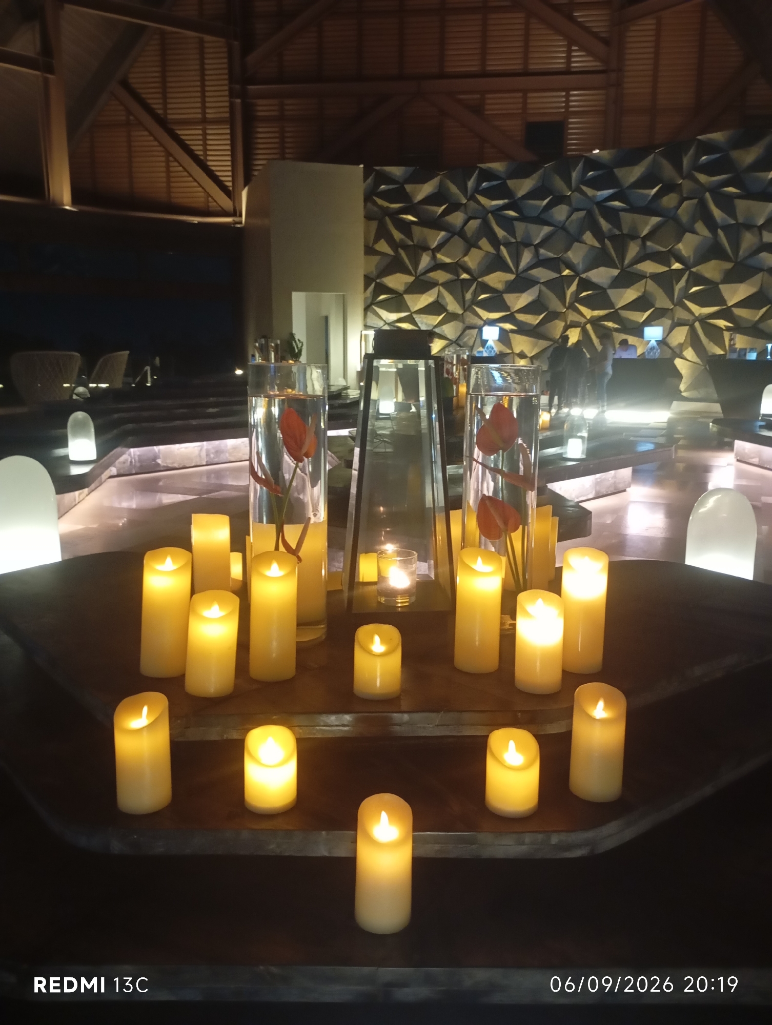 The image depicts a dimly lit interior space, likely a hotel lobby or lounge, with an emphasis on decorative lighting and ambiance. In the foreground, a collection of pillar candles of varying heights are arranged on a dark, circular surface. The candles are illuminated, emitting a warm, yellowish glow. Behind the candles, a tall glass vase contains water and two red calla lilies. Further back, a unique geometric wall feature is visible, illuminated from behind, creating a textured and patterned backdrop. In the background, several people are present, some appearing to be staff at a counter, while others are seated or standing, suggesting a reception or waiting area. The overall atmosphere is one of luxury and tranquility. The image was taken in Kuta Selatan, Indonesia, as indicated by the timestamp.