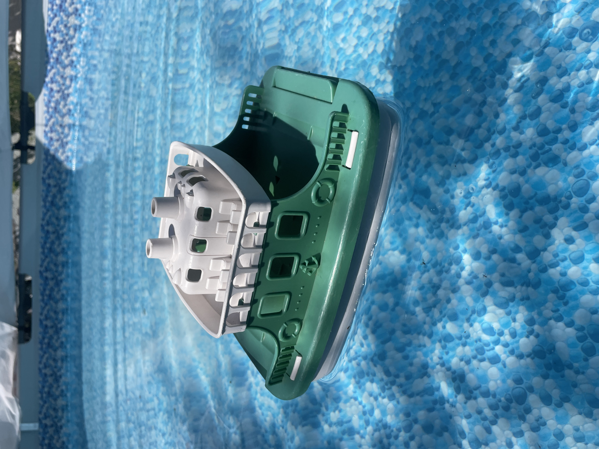 A toy boat, predominantly green with a white upper deck, floats in the shallow, clear blue water of a swimming pool. The bottom of the pool is lined with a pattern of blue and white circular shapes. The boat has several details, including windows, a life preserver, and railing. The top of the boat is equipped with two smokestacks and multiple seating areas. Sunlight reflects off the water's surface, creating ripples and bright highlights. The scene is set in an outdoor environment, likely a backyard, given the presence of the swimming pool. There are no people visible in the image.