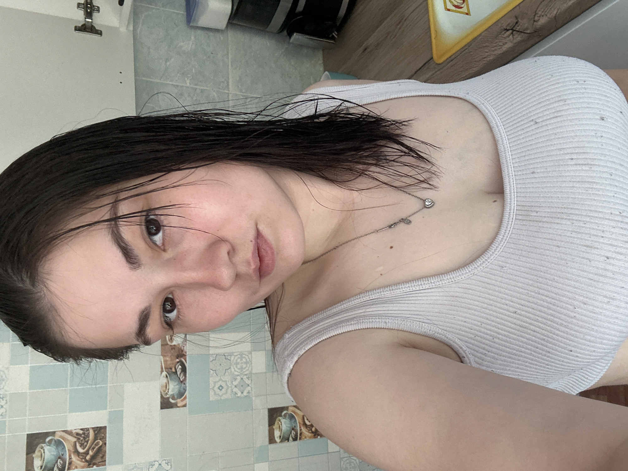 A young woman with dark, wet hair is taking a selfie in what appears to be a kitchen. She is wearing a light-colored ribbed tank top and a silver necklace with a heart-shaped pendant. The background shows a tiled wall with a coffee cup design, a wooden countertop, and kitchen appliances. The lighting suggests it is daytime. There is no visible indication that this scene is located in Chaikovsky, Russia.