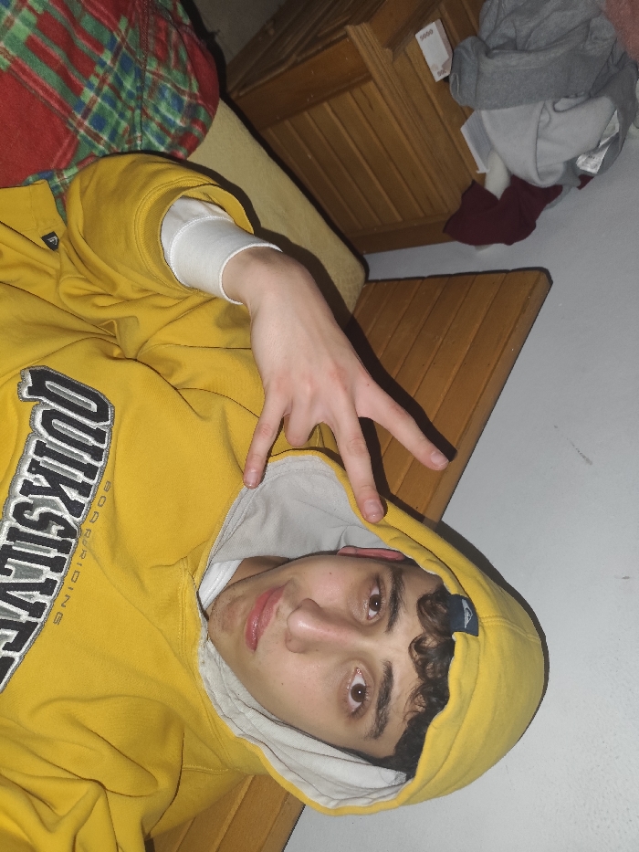 A young man wearing a yellow "Quiksilver Boardriding" hooded sweatshirt and a white undershirt lies on a surface with a wooden texture. He is making a hand gesture with his right hand. His head is tilted, and he is looking directly at the camera. Behind him, there's a wooden dresser and some rumpled clothing. The wall behind the dresser appears to be white. Part of a plaid blanket or fabric is visible in the upper left corner.