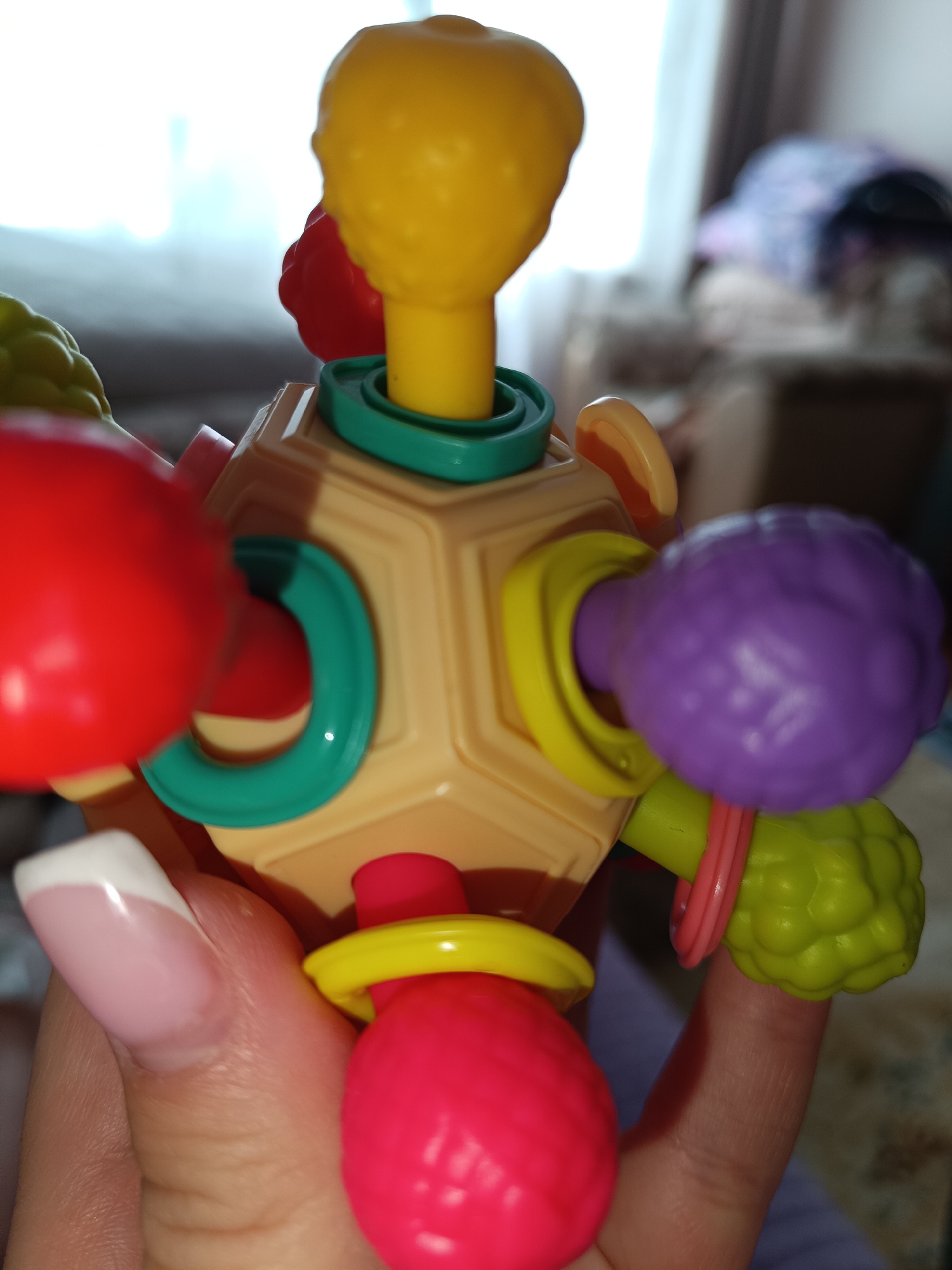 A person's hand holds a multifaceted, plastic children's toy with nubby, colorful spheres protruding from the faces. The toy is primarily tan with green and yellow rings around the sphere stems. The spheres are red, yellow, purple, and lime green. The hand has a manicured nail with a French tip design. The background is blurry, but contains a window and furniture. The location is not explicitly specified in Tobolsk, Russia.
