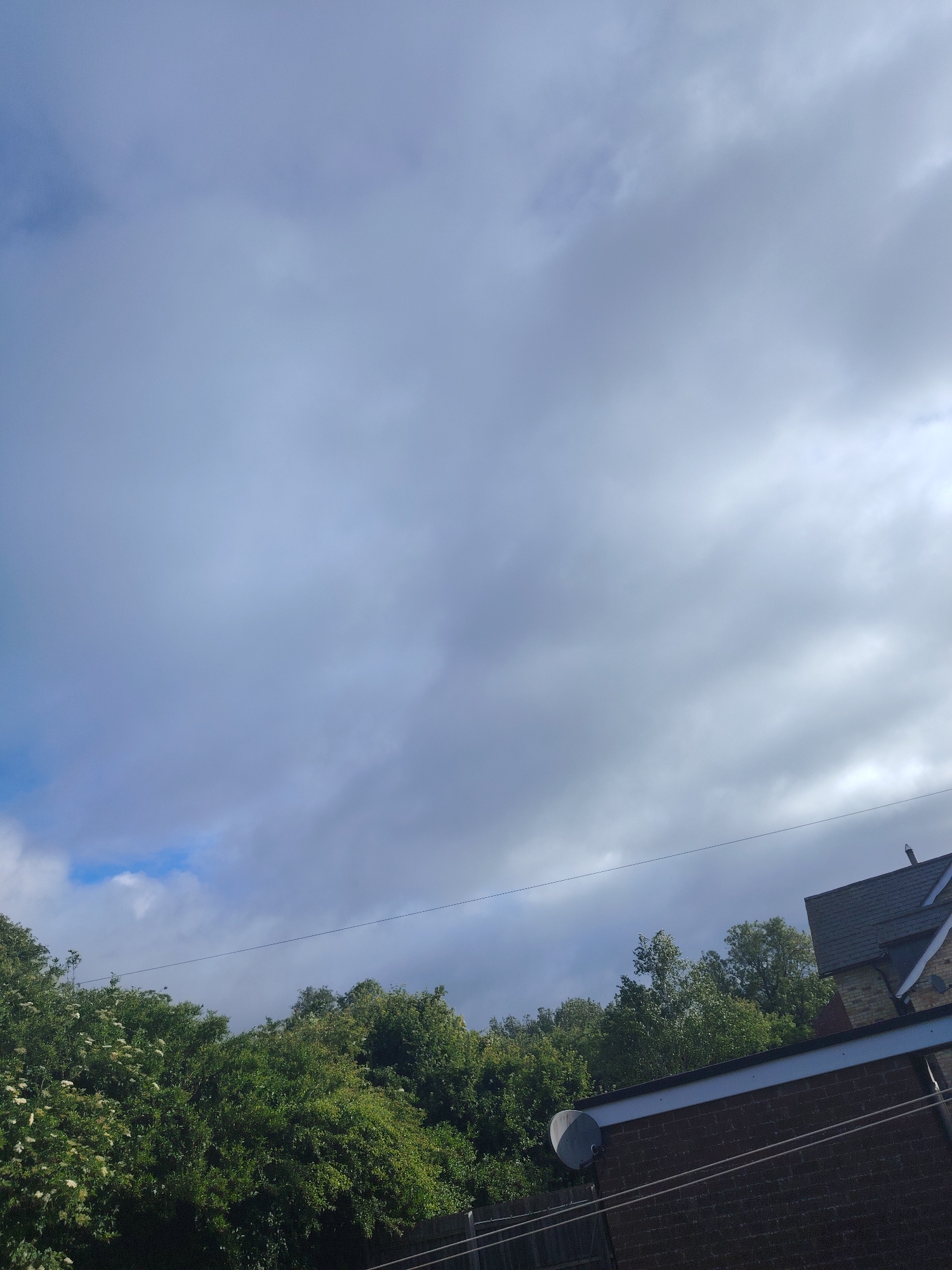 The image captures an outdoor scene dominated by a mostly cloudy sky, with patches of clear blue visible in the lower-left and upper-left sections. The clouds are a mix of light grey and white, suggesting diffused daylight. A thin, dark cable or power line stretches horizontally across the middle of the frame, just above a dense band of green trees and bushes that spans the lower portion of the image. Some white blossoms are visible within the foliage on the left side.
On the right, the upper part of a brick building is visible, featuring a dark grey tiled roof with a prominent chimney structure. Below the main roofline, a section of flat roof with a white fascia is present, and a grey satellite dish is mounted on the brick wall beneath it. Another dark horizontal line, possibly a fence, runs along the very bottom edge of the image.
No people or specific activities are depicted. The weather appears to be partly cloudy to overcast during daytime hours. The architectural style and natural elements are consistent with a suburban or semi-rural environment, typical of areas like Huntingdonshire, United Kingdom. There is no visible text.