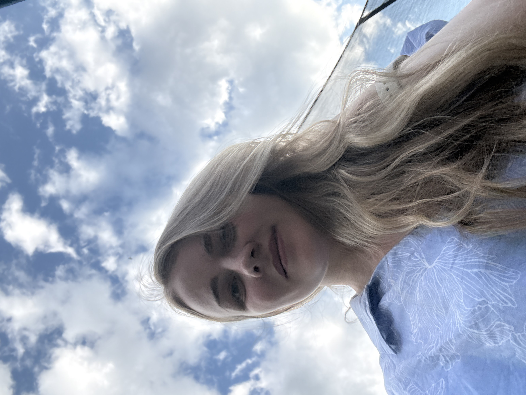 A low-angle shot depicts a woman with long blonde hair, wearing a light blue floral-patterned shirt. The background features a sky with scattered clouds. There is no specific information present to indicate that this image was taken in the city of Chaikovsky, Russia. The subject appears to be looking down at the camera.