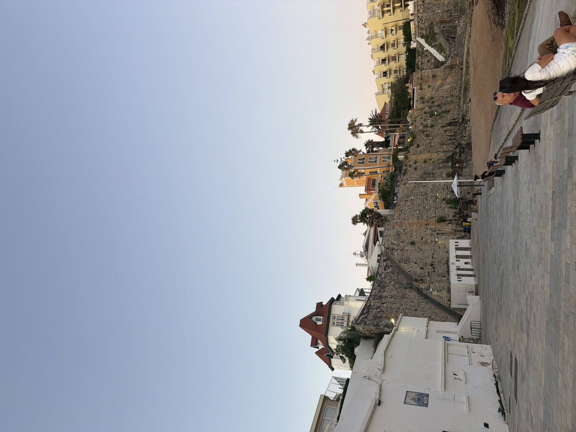 This image captures a view of the coastline in Cascais, Portugal, likely near the Praia da Rainha or a similar beach area. The scene is set during what appears to be late afternoon or early evening, with the sky a clear, pale blue, transitioning to a softer hue at the horizon.
In the foreground, a paved promenade runs along the edge of a sandy beach. Benches are arranged along the promenade, with a few individuals seated on them, enjoying the view. To the left, a white building with a blue tile plaque is visible, featuring windows and a slightly weathered facade. Further along the promenade, a row of small structures, possibly public restrooms or changing rooms, are lined up against a stone retaining wall.
Dominating the middle ground is a dramatic cliff face, constructed from rough-hewn stones. A set of stairs ascends this wall, leading to buildings perched at the top. These buildings include a white structure with a distinctive red-tiled roof, featuring multiple gables and dormer windows, and a tall, bright yellow building with arched windows and balconies, reminiscent of a hotel or large residence. Palm trees are scattered throughout this upper section, adding to the coastal ambiance. In the background, more yellow buildings with balconies can be seen, suggesting a developed coastal town.
Two people are the most prominent subjects in the foreground, seated on a bench facing away from the camera. A man with gray hair and sunglasses is wearing a maroon shirt, while a woman beside him has long dark hair and is wearing a white top and patterned shorts. They appear to be in conversation, looking towards the sea. Other figures are more distant and less distinct, scattered along the promenade and on the beach. The overall impression is one of a peaceful seaside setting, with a blend of natural landscape and architectural elements.