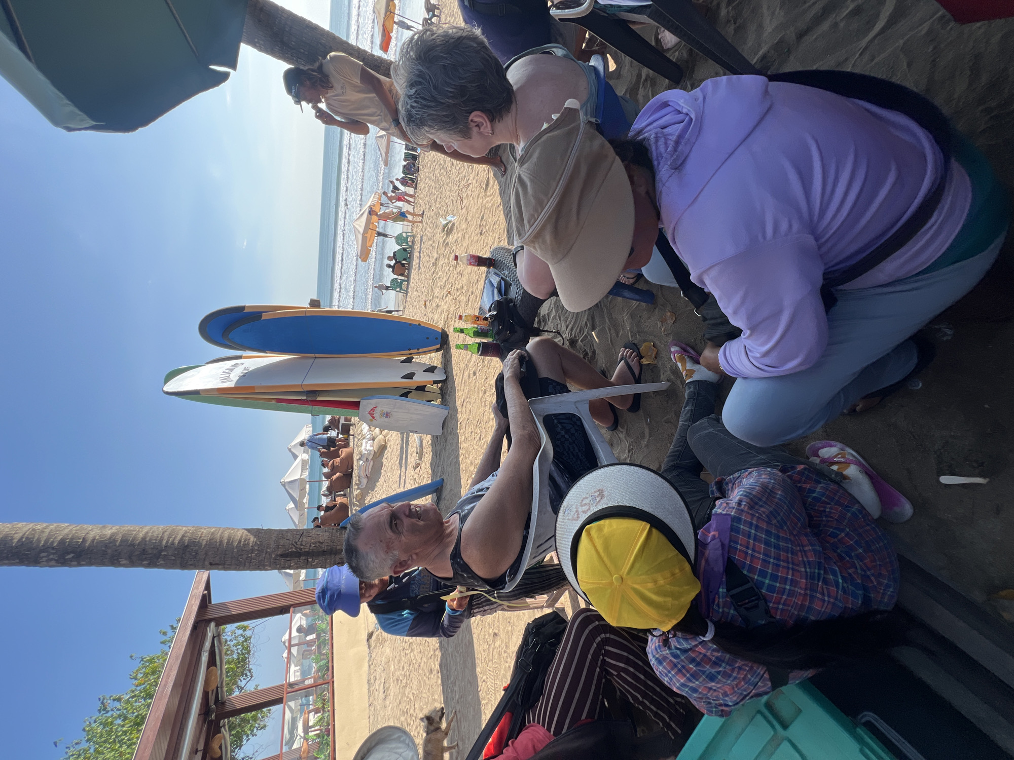 In Kuta, Indonesia, on a sunny beach, a group of people are gathered in front of a row of surfboards. A man with gray hair and a black tank top sits in a white chair, facing right, appearing to speak with someone out of frame. Next to him, a person wearing a blue hat and jacket stands, holding a yellow object. In the foreground, a person in a yellow hat and a plaid shirt sits on the sand, their back to the camera. To their right, a person in a purple hoodie and light blue pants is also seated. Behind them, another person in a light blue top and a wide-brimmed hat is visible. A table with drinks and bottles sits between the man in the chair and the people in the foreground. In the background, the ocean, beach umbrellas, and other beachgoers are visible under a clear blue sky.