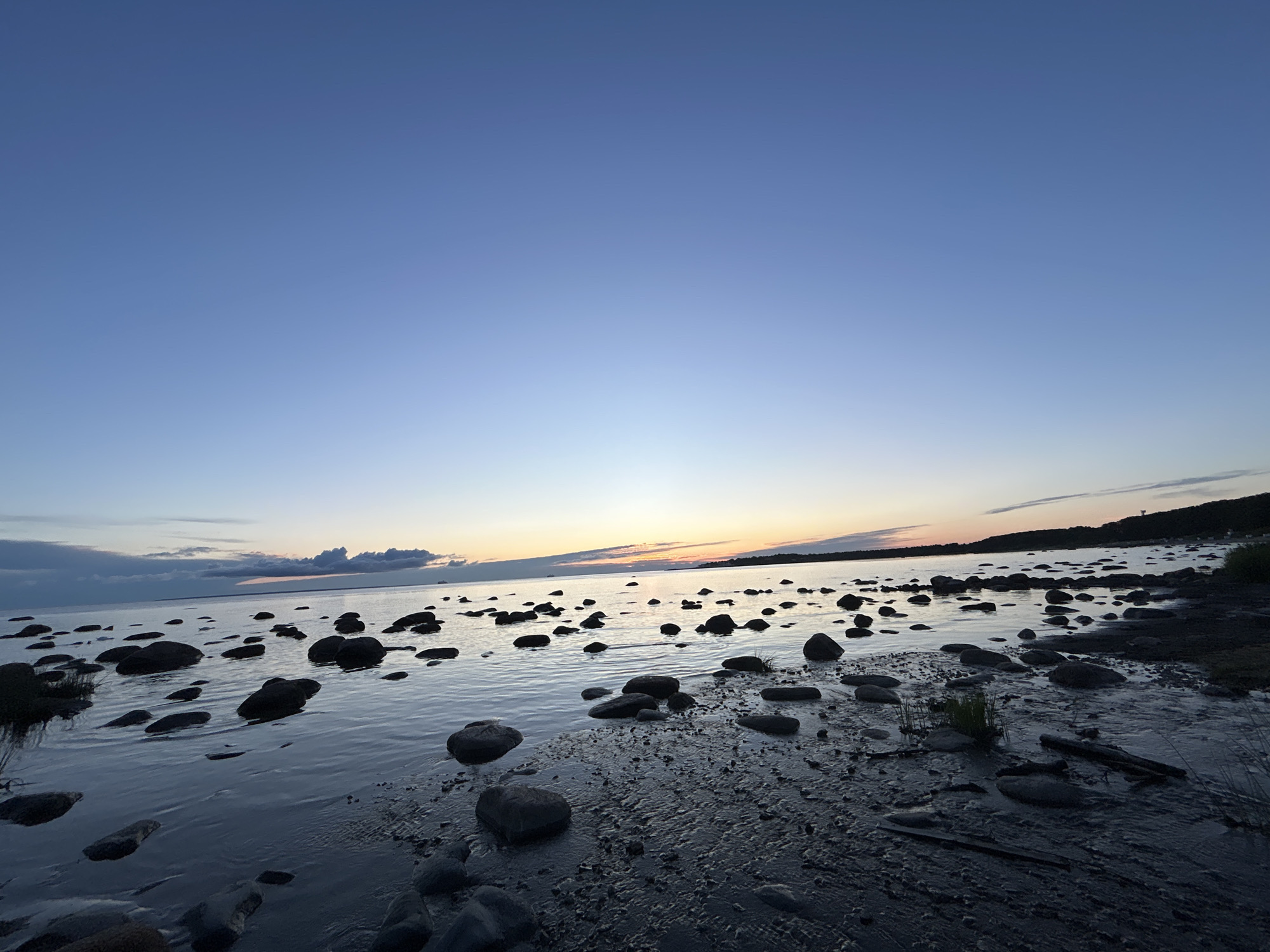 The image depicts a rocky shoreline at dusk in Haabneeme alevik, Estonia. The sky is a gradient of deep blue to light blue, with a faint orange and yellow glow on the horizon. Scattered across the dark, wet sand and shallow water are numerous dark, smooth rocks of varying sizes. The water reflects the colors of the sky. In the distance, a silhouetted landmass with trees and a small structure is visible. There are no people or animals in the frame. The overall scene is serene and natural.