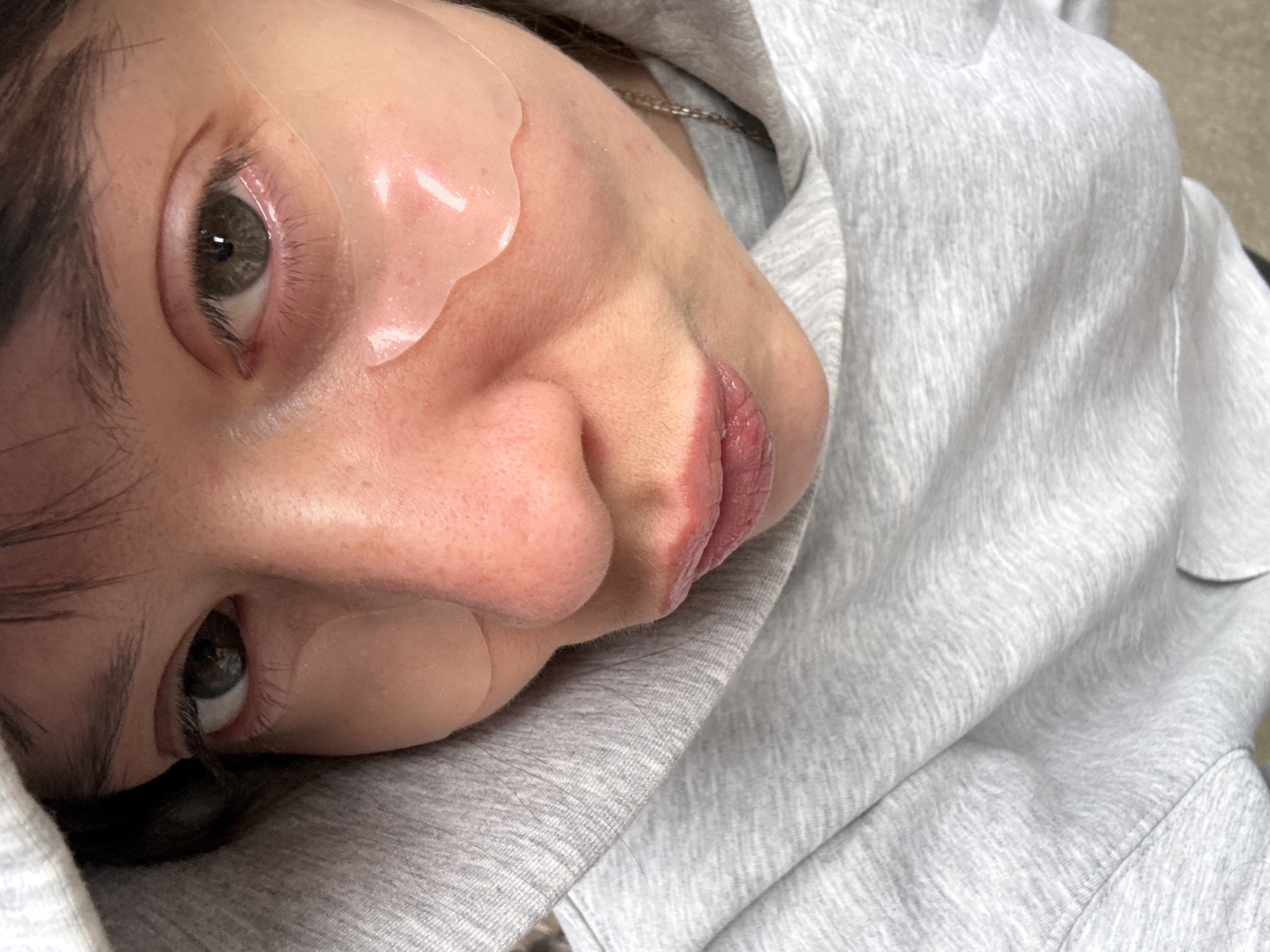 This close-up, angled image captures a person, likely female, from a slightly elevated perspective as they lie on their side. The subject has medium-toned skin, dark hair, and striking green or hazel eyes that are open and looking towards the viewer. Their lips are naturally pink and slightly parted, appearing moisturized or glossy.
A prominent feature is the application of clear, gel-like under-eye patches, one visible beneath each eye, reflecting ambient light and suggesting a skincare or beauty treatment. The person is wearing a light grey hooded sweatshirt, with the textured fabric visible around their head and shoulders, implying a casual and comfortable setting. A silver chain is partially visible around their neck, peeking out from beneath the hood.
The activity appears to be a moment of relaxation or self-care. The subject's expression is neutral and calm. The lighting is soft and even, highlighting the contours of the face and the texture of the skin. The background is out of focus, providing no specific location cues, though the provided context indicates London, United Kingdom. There is no visible text in the image.