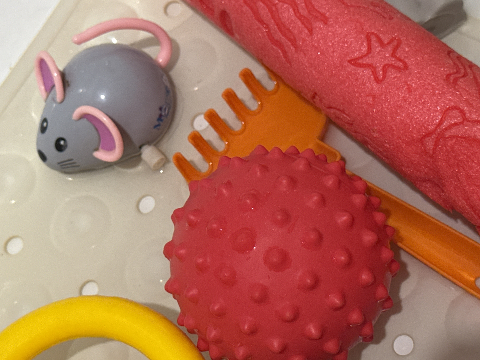 In a close-up, high-angle shot, a collection of children's bath toys rests on a textured, light-colored surface that appears to be a bathtub or shower floor. The toys include a bright yellow ring, a textured red ball, an orange toy rake, a wind-up gray mouse with pink ears and tail, and a red foam roller with embossed shapes of stars and possibly waves. Water droplets are visible on some of the toys, suggesting they are wet. The scene is well-lit, and the focus is sharp on the toys. The specific location in Яромінскі сельскі Савет, Belarus, is not discernible from the visual information presented in the image.