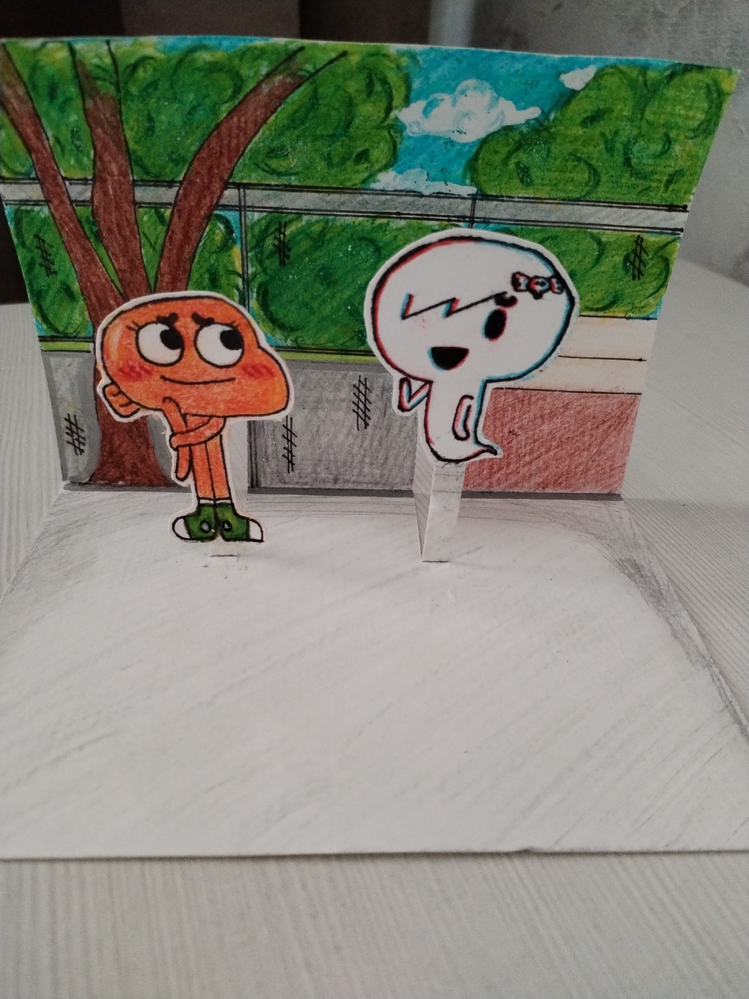 This is a close-up, eye-level, full shot of a handmade diorama. The diorama depicts a stylized outdoor scene. In the foreground, two cartoon characters are depicted as stand-up figures. The character on the left is orange and round with large, expressive eyes and green shoes. The character on the right is white, ghost-like, with black eyes and a bow on its head. The background of the diorama is a painted scene of green trees, blue sky with white clouds, and a fence. The diorama is set on a white, textured surface. There is no discernible information about the location in Хідрынскі сельскі Савет, Belarus.