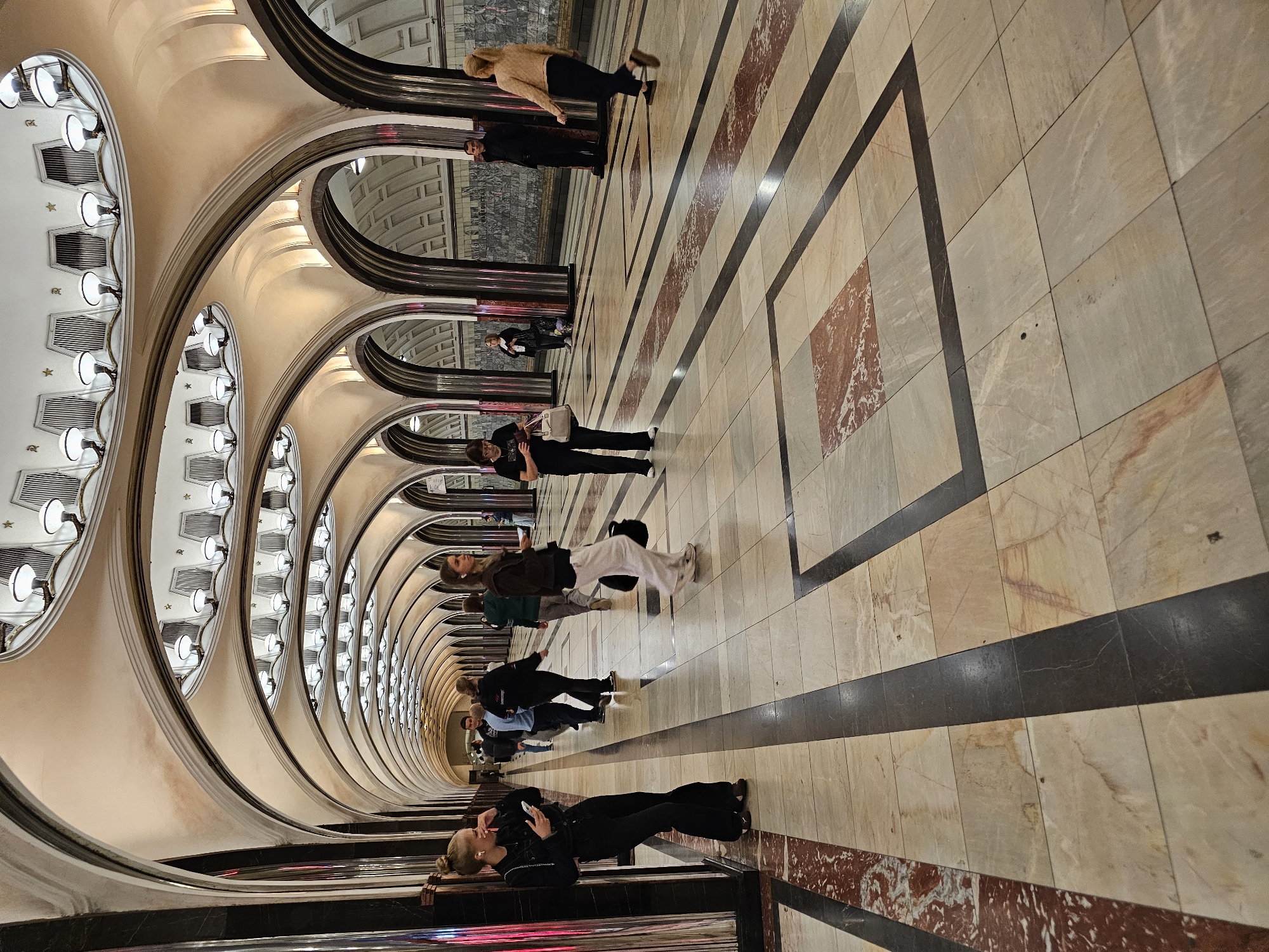 The image shows a view down a long, arched hallway of a Moscow Metro station, likely Komsomolskaya station due to its ornate architecture. The hallway is lined with marble columns and features a high, vaulted ceiling with elaborate light fixtures. The floor is paved with polished stone tiles in a geometric pattern, with darker stripes. Several people are walking in both directions along the hallway. In the foreground on the left, a young woman in black is leaning against a column, looking at her phone. Further down, other individuals are visible, some walking and some standing. The walls of the station are adorned with mosaic panels, partially visible through arched openings. The overall impression is one of grandeur and spaciousness, characteristic of Moscow's famous Metro system.