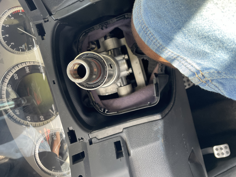 The image depicts the interior of a vehicle from the driver's perspective. The specific location in Alaiyabiagba, Nigeria, is not discernible from the image. The steering wheel is absent, exposing the silver-colored, threaded steering column shaft and associated mechanical components, surrounded by black trim and purple-grey insulation material. Above, the vehicle's instrument cluster is visible, showing a speedometer peaking at 160 units and a tachometer up to 7,000 RPM, beneath a clear protective cover. To the lower left, a metallic, perforated pedal is visible. In the lower right quadrant, a person's right leg, clad in blue denim, is present in the footwell area. The scene indicates the vehicle is in a state of disassembly or repair.