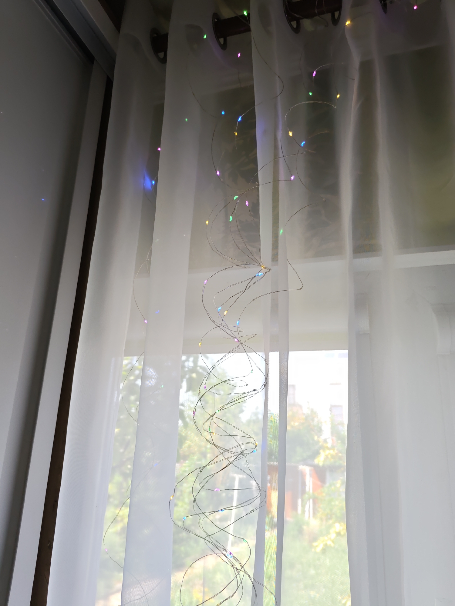 The image shows a sheer white curtain hanging in a window, adorned with a string of colorful fairy lights. The lights are small, LED bulbs in various colors, including pink, blue, green, and yellow, which are attached to a thin, wire-like string. The string is draped loosely over the curtain, creating a whimsical, decorative effect. Through the sheer curtain, a blurred outdoor scene is visible, with hints of trees and buildings bathed in natural light, suggesting a daytime setting. The overall impression is one of a softly lit interior space, perhaps a living room or bedroom, enhanced by the festive glow of the fairy lights. The specific location of Rostov-on-Don, Russia, cannot be determined from the visual information provided in the image.