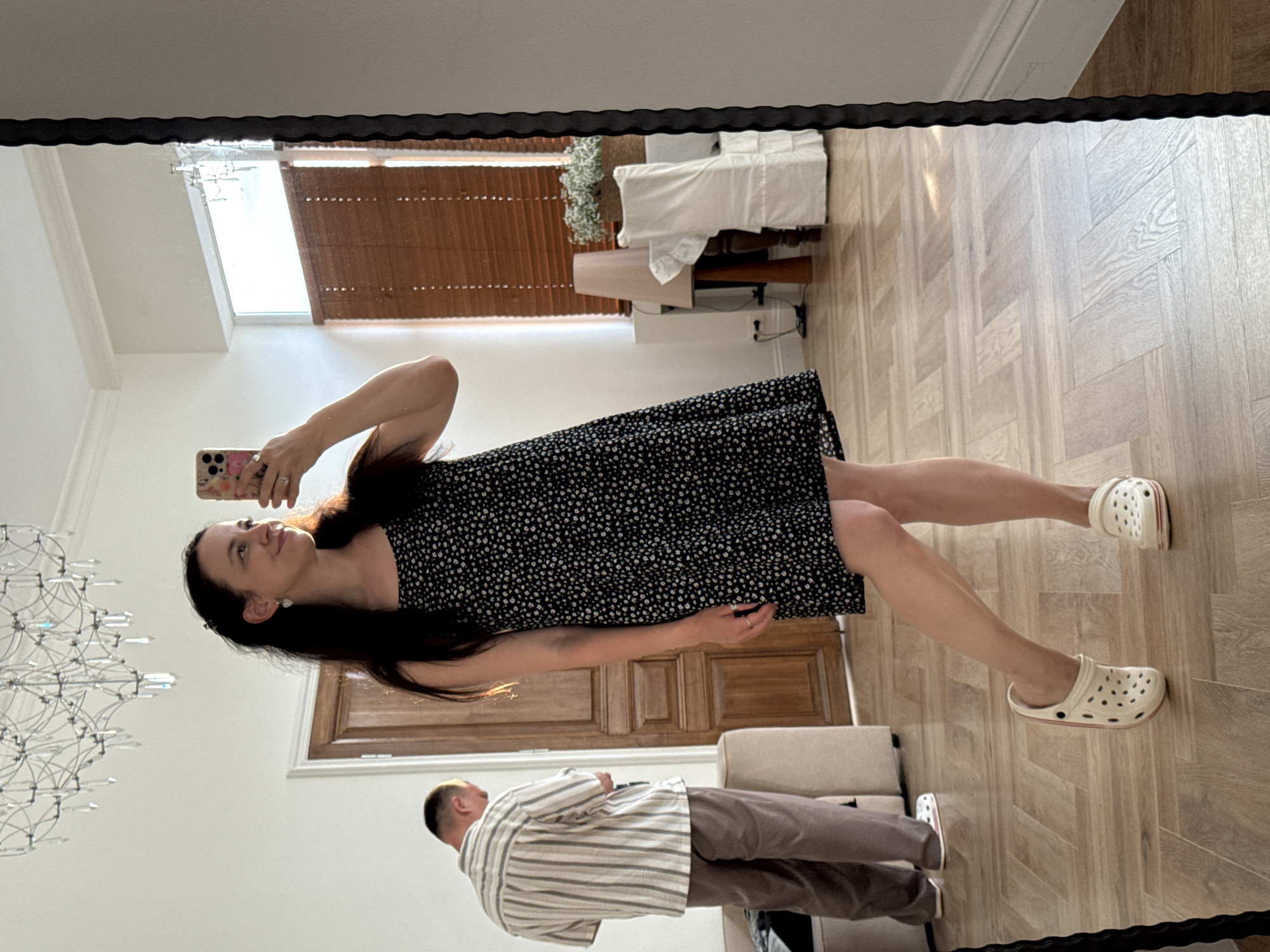 This media file captures a reflection in a mirror, showcasing two individuals and an indoor setting. The primary subject is a woman in the foreground, taking a mirror selfie with her smartphone. She has long, dark hair and is wearing a black dress with a white speckled pattern and white clogs. Her right arm is raised, holding the phone, and she is looking up and to the side with a relaxed expression.
Behind her, and also reflected in the mirror, is a man wearing a striped collared shirt and brown trousers. He is facing away from the camera, looking down. The setting appears to be a well-lit room with light wood-paneled flooring arranged in a herringbone pattern. Visible in the reflection are wooden doors and a large, ornate chandelier on the left. The background also shows a window with horizontal blinds, suggesting natural light is entering the room. The overall mood is casual and domestic. There is no visible text in the image.