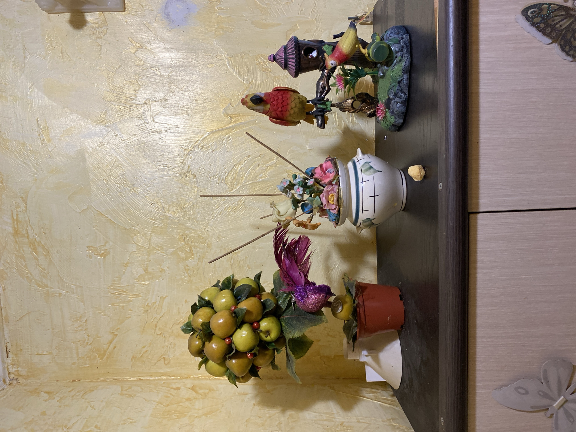 The image depicts a shelf in what appears to be a residential setting, likely a home in Maykop, Russia. The wall behind the shelf has a textured, yellow-gold finish.
On the shelf, there are several decorative items. On the left is a potted artificial tree adorned with green leaves and yellow fruits, with a small purple bird figurine attached to its trunk. Next to it is a white ceramic vase filled with artificial flowers and several incense sticks. To the right, there is a decorative piece featuring a birdhouse with two colorful bird figurines. One bird is perched on the birdhouse roof, and another is on a branch in front of it. A small, light-colored crumb or pellet is also visible on the shelf.
The shelf itself is dark brown and appears to be the top of a piece of furniture, possibly a cabinet or dresser, which has light-colored doors with butterfly decals on them. The lighting in the scene is relatively soft, casting some shadows. The overall impression is of a decorated shelf within a domestic environment.