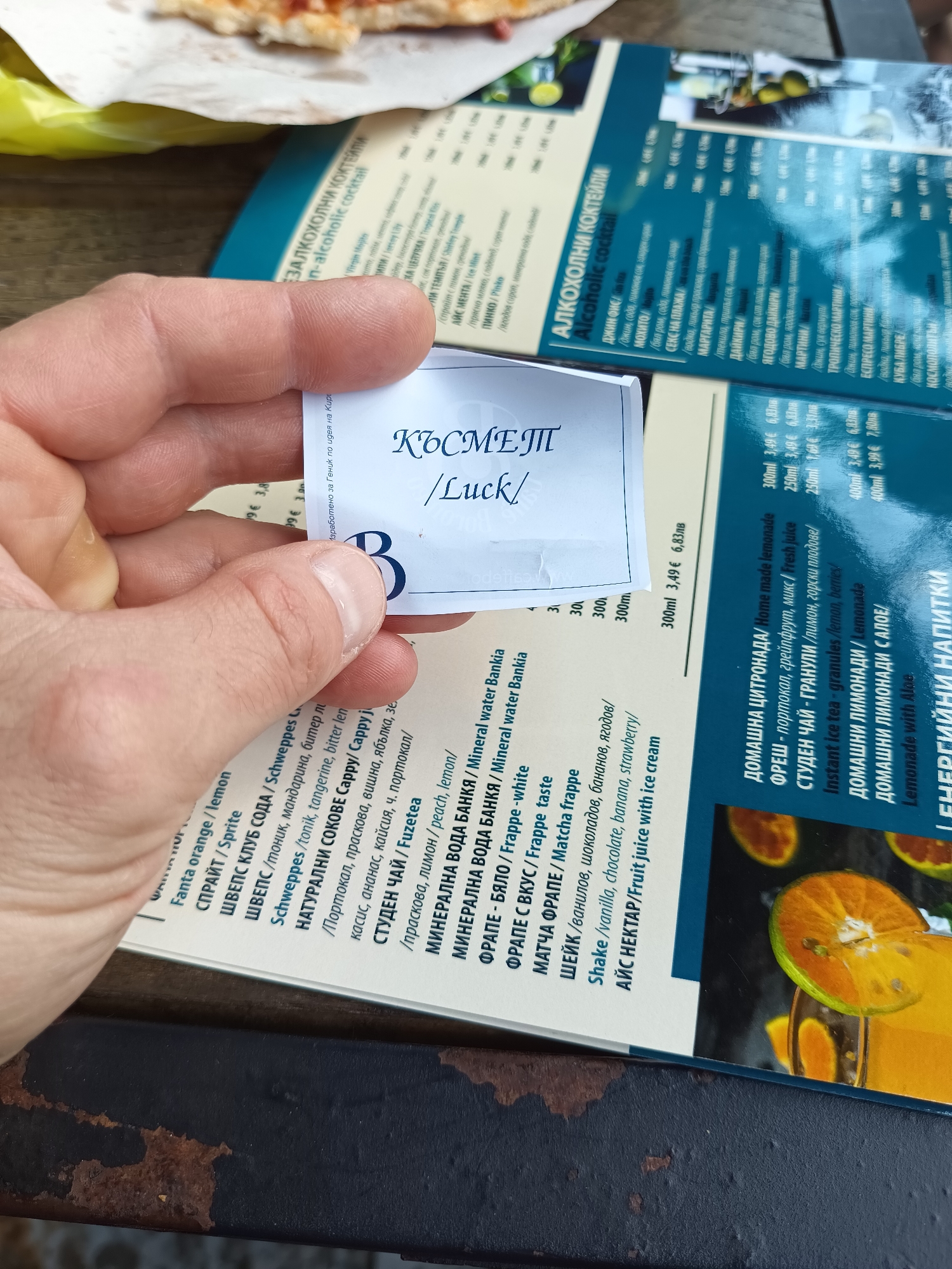 The image captures a close-up scene, likely in an outdoor dining setting. A person's hand is holding a small paper card with "КЪСМЕТ/Luck/" printed on it, along with the letter "B". Beneath the card, part of a menu is visible, featuring Bulgarian text listing various drinks, including non-alcoholic cocktails, soft drinks, and alcoholic cocktails. The menu lists prices and descriptions in both Bulgarian and English. In the background, there are hints of food (possibly pizza) and other menu sections. The scene is most likely at an outdoor restaurant or café in Targovishte, Bulgaria given that the text is in Bulgarian, and "Геник" is from Targovishte.